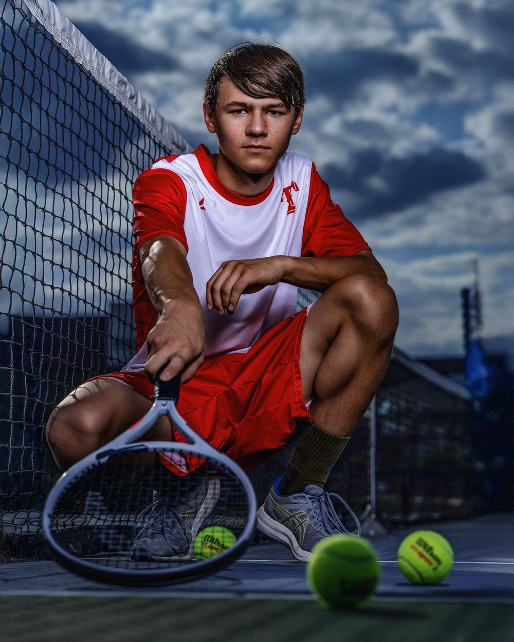 Dramatic portrait of a tennis player crouching on court with racquet and balls against stormy sky and net background.