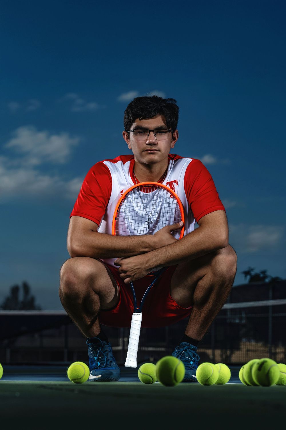 Tennis player in red and white uniform crouches on court with tennis racket and balls during outdoor photo session.