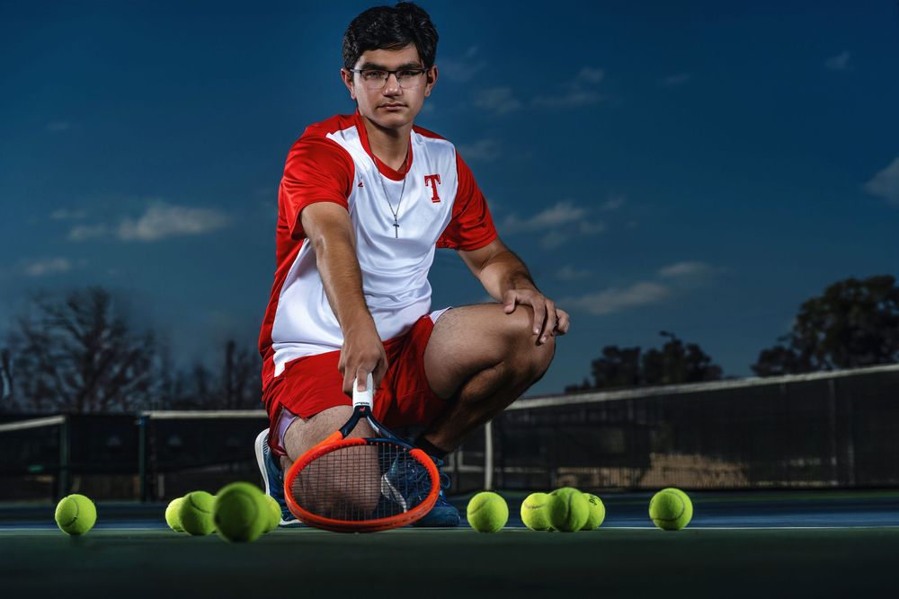 Tennis player in red and white uniform crouches on court with tennis balls and racquet during dramatic evening photoshoot.