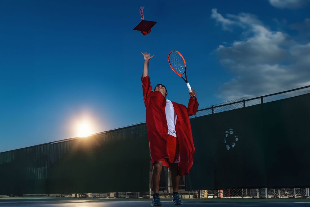 Graduate in red cap and gown tosses their cap while celebrating outdoors during sunset with tennis racquet in hand.