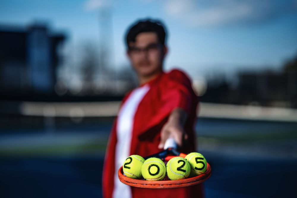 Tennis balls in a yellow holder shown with blurred figure in red athletic wear in background on outdoor tennis court.