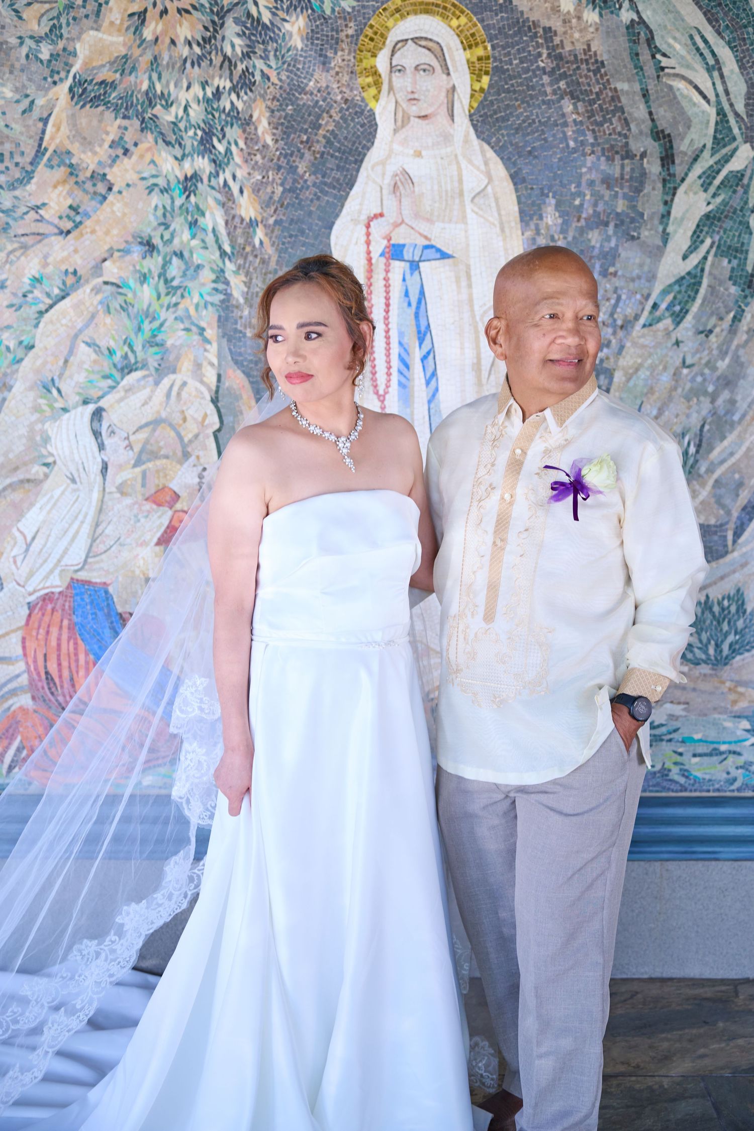 A wedding couple poses against a religious mural backdrop with flowing white veil and dress at a church.