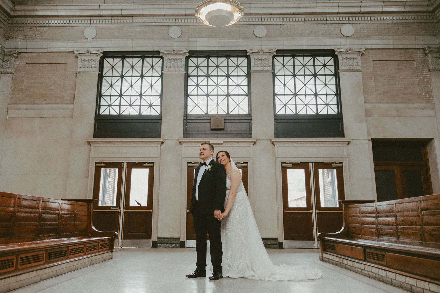 A couple poses in formal wedding attire inside a grand historic building with wooden pews and architectural windows.