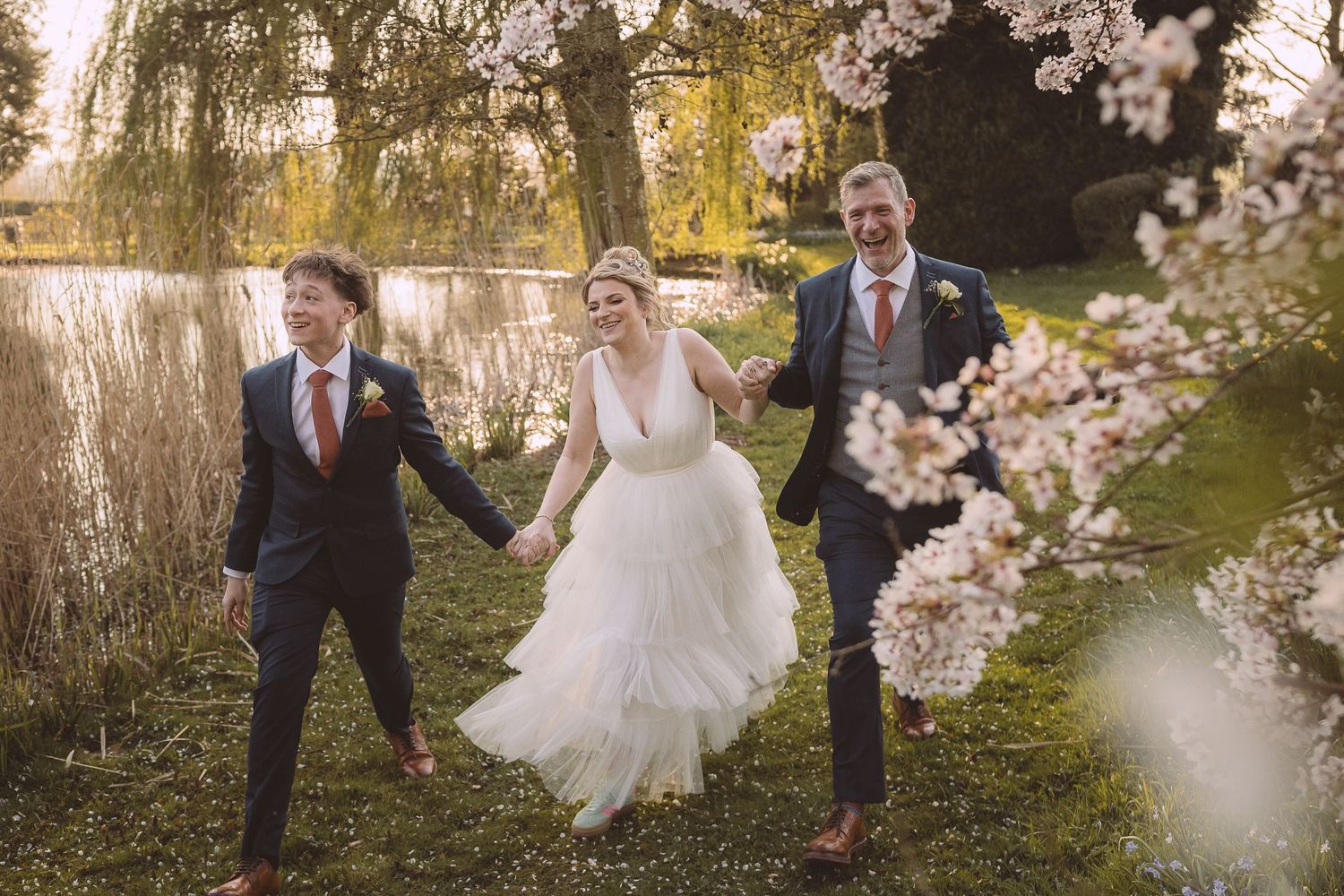Three people run joyfully through a garden with cherry blossoms and reeds beside a pond on a sunny spring day.