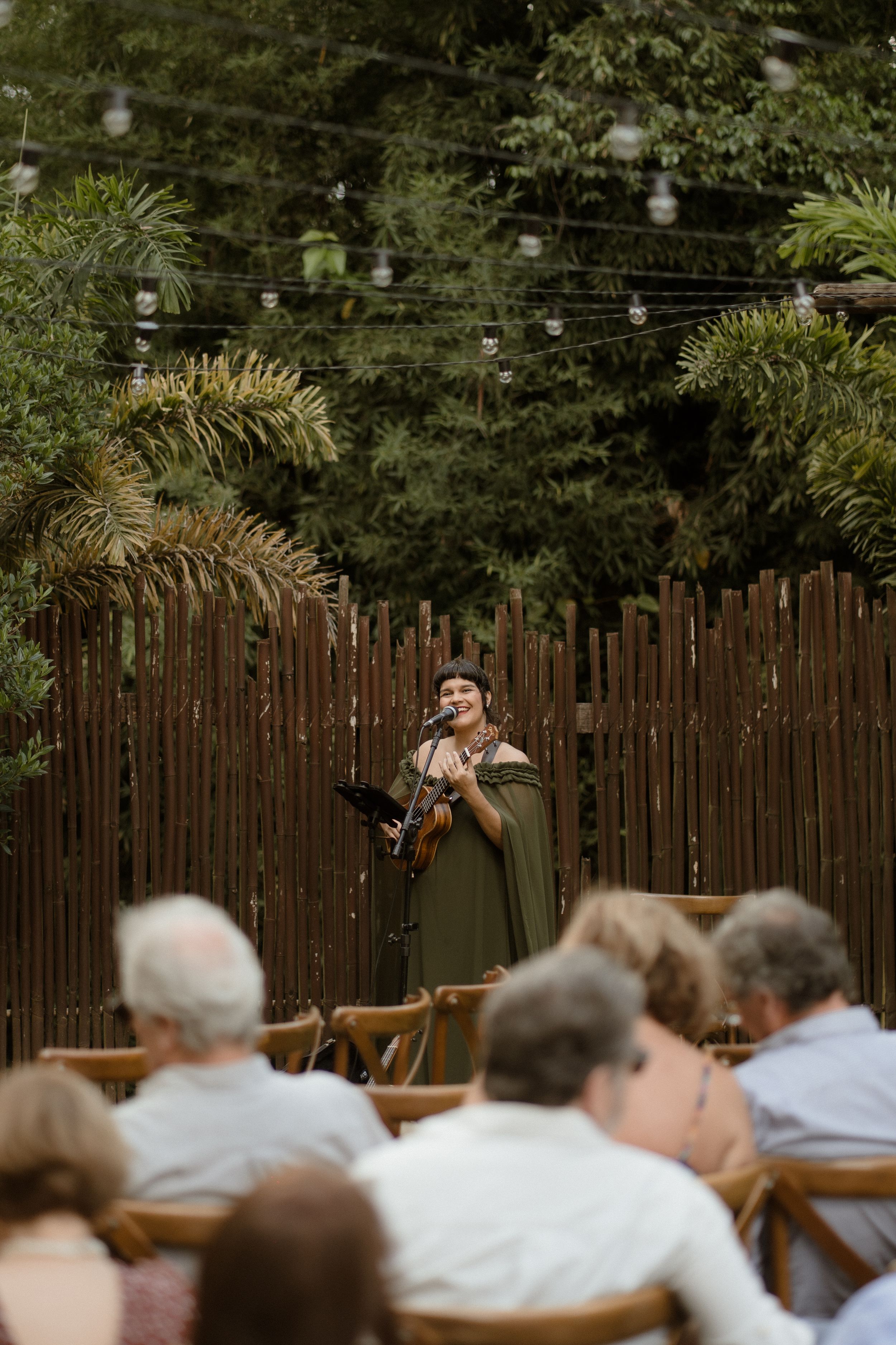 Musician performing at an outdoor venue with string lights and wooden fence backdrop.