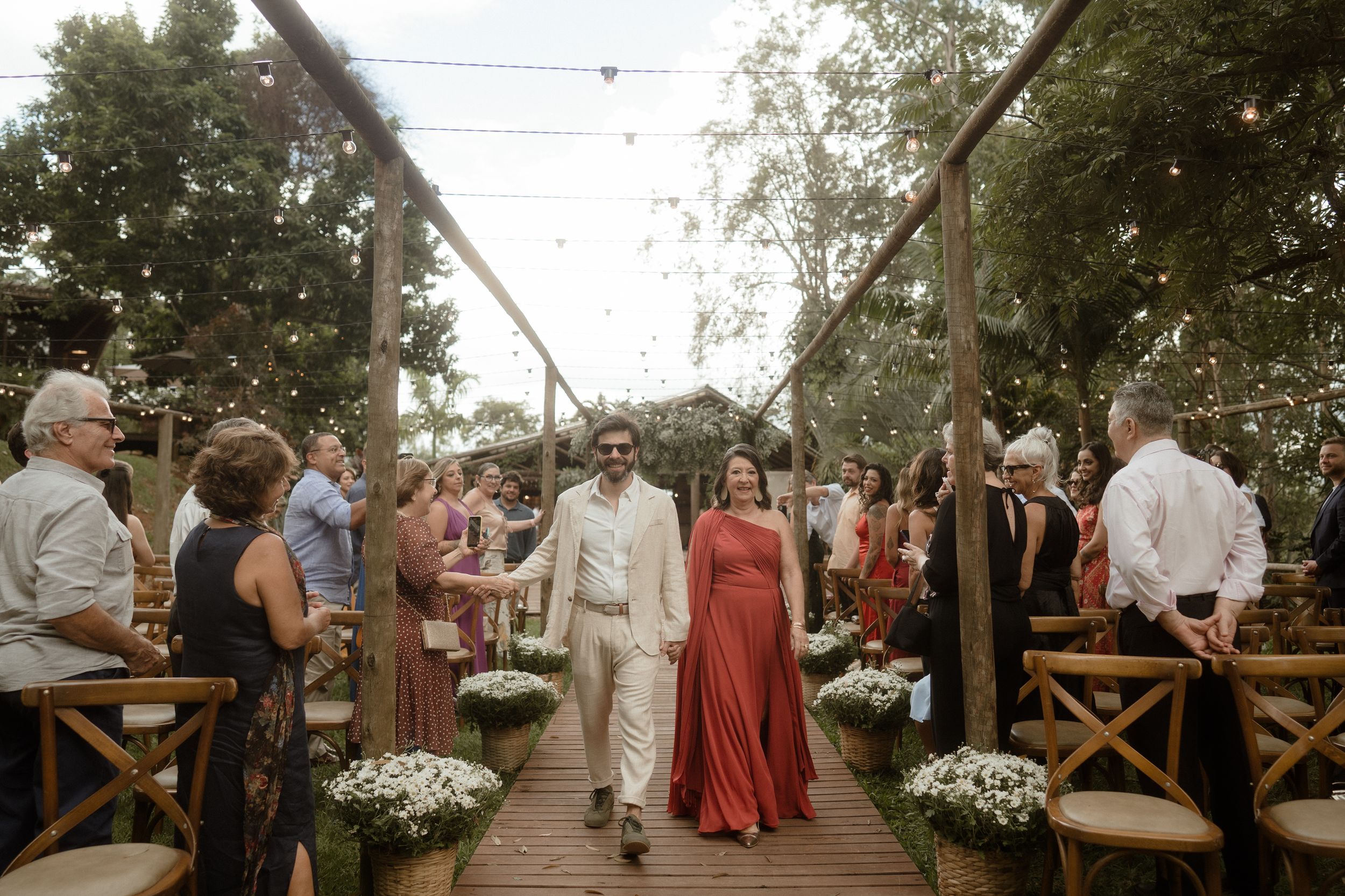 Wedding guests seated in wooden chairs along an outdoor aisle decorated with white flowers.