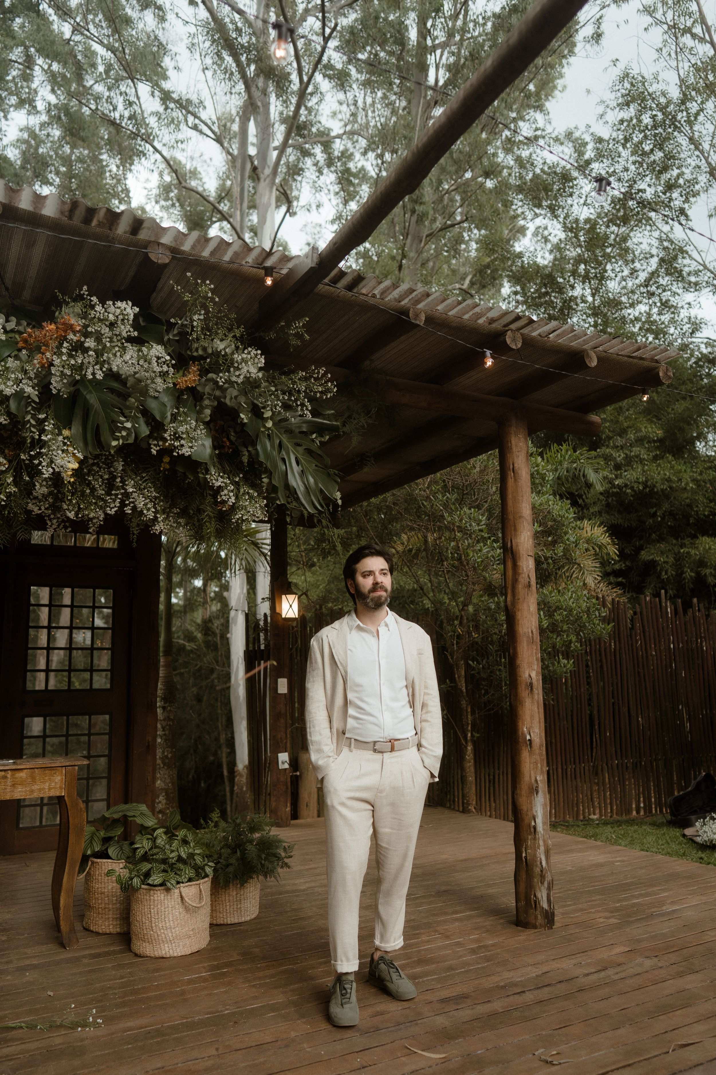 An outdoor wedding venue with wooden pergola, string lights, and rustic decor against a backdrop of tall trees.