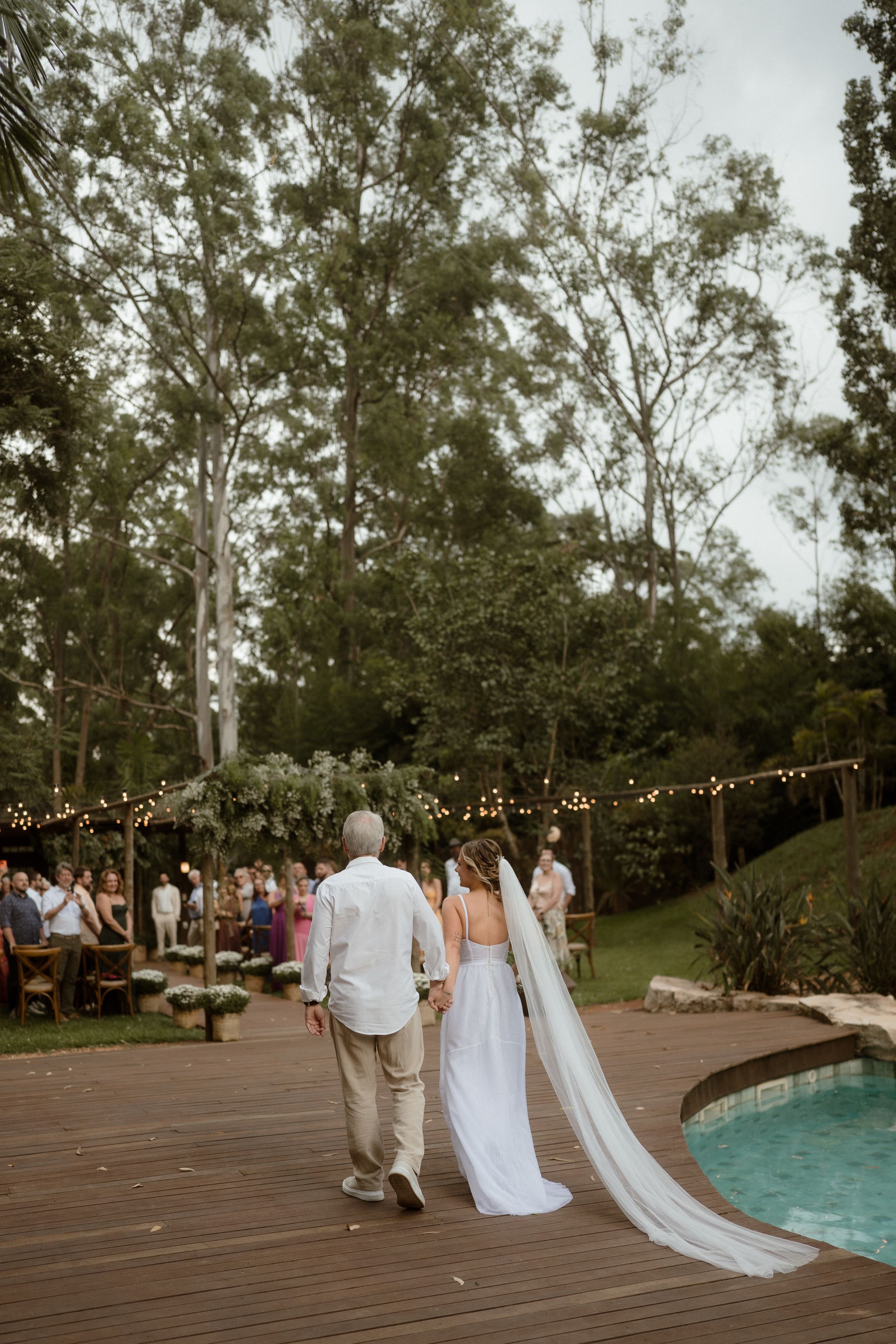 A couple walking near a pool at an outdoor wedding with string lights and guests in the background.
