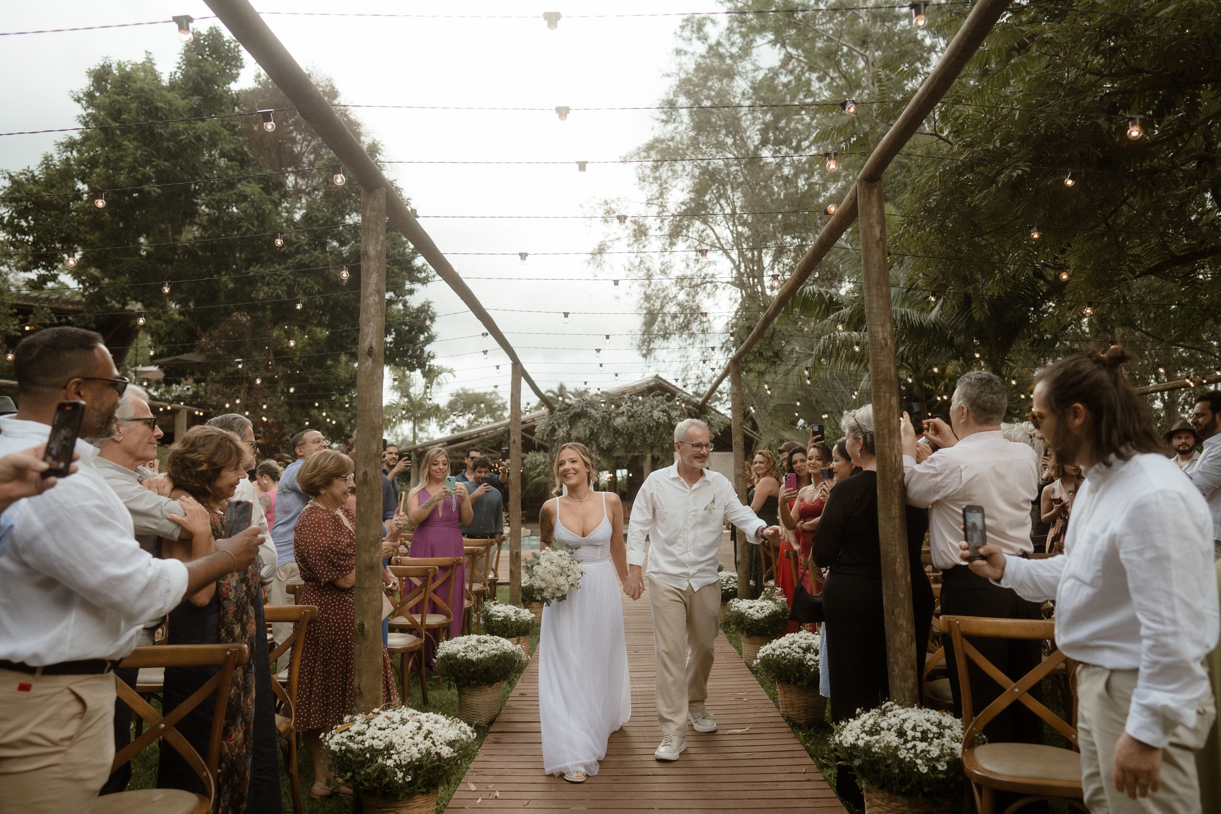 Wedding guests line a wooden walkway under string lights as newlyweds walk between them.