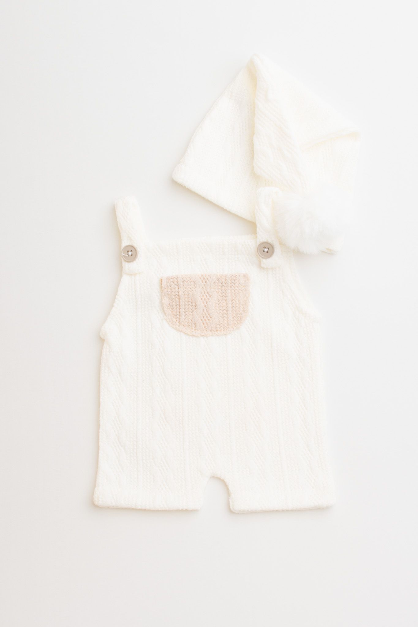 White knit baby overalls with tan chest pocket and button straps on neutral background.