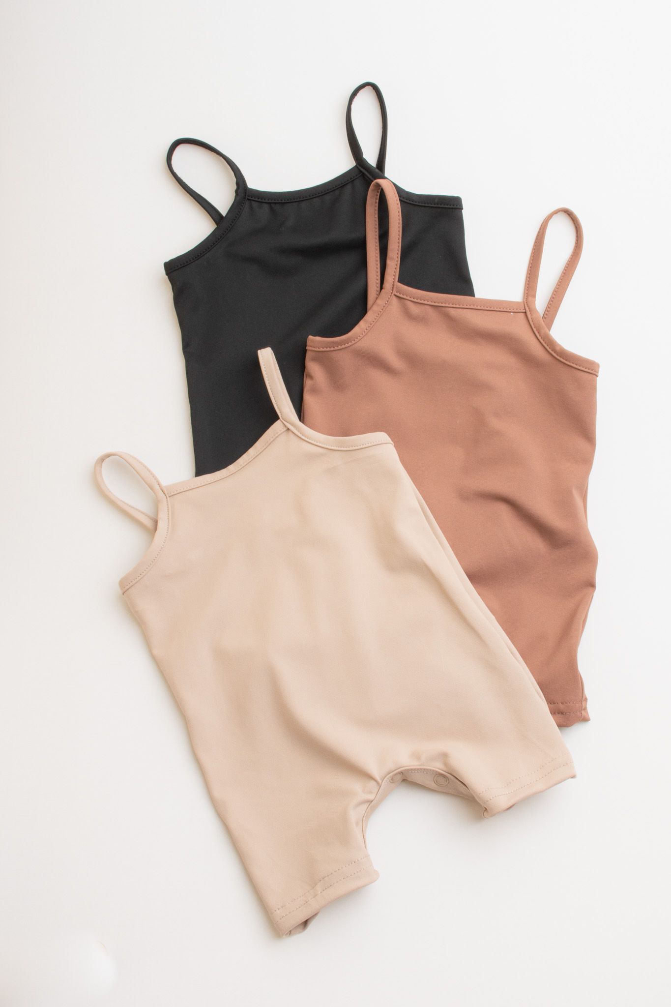 Three baby rompers in neutral colors black, beige and brown laid flat on white background.