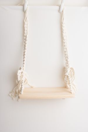 Macrame swing with wooden seat and white braided ropes hanging against a plain white wall.