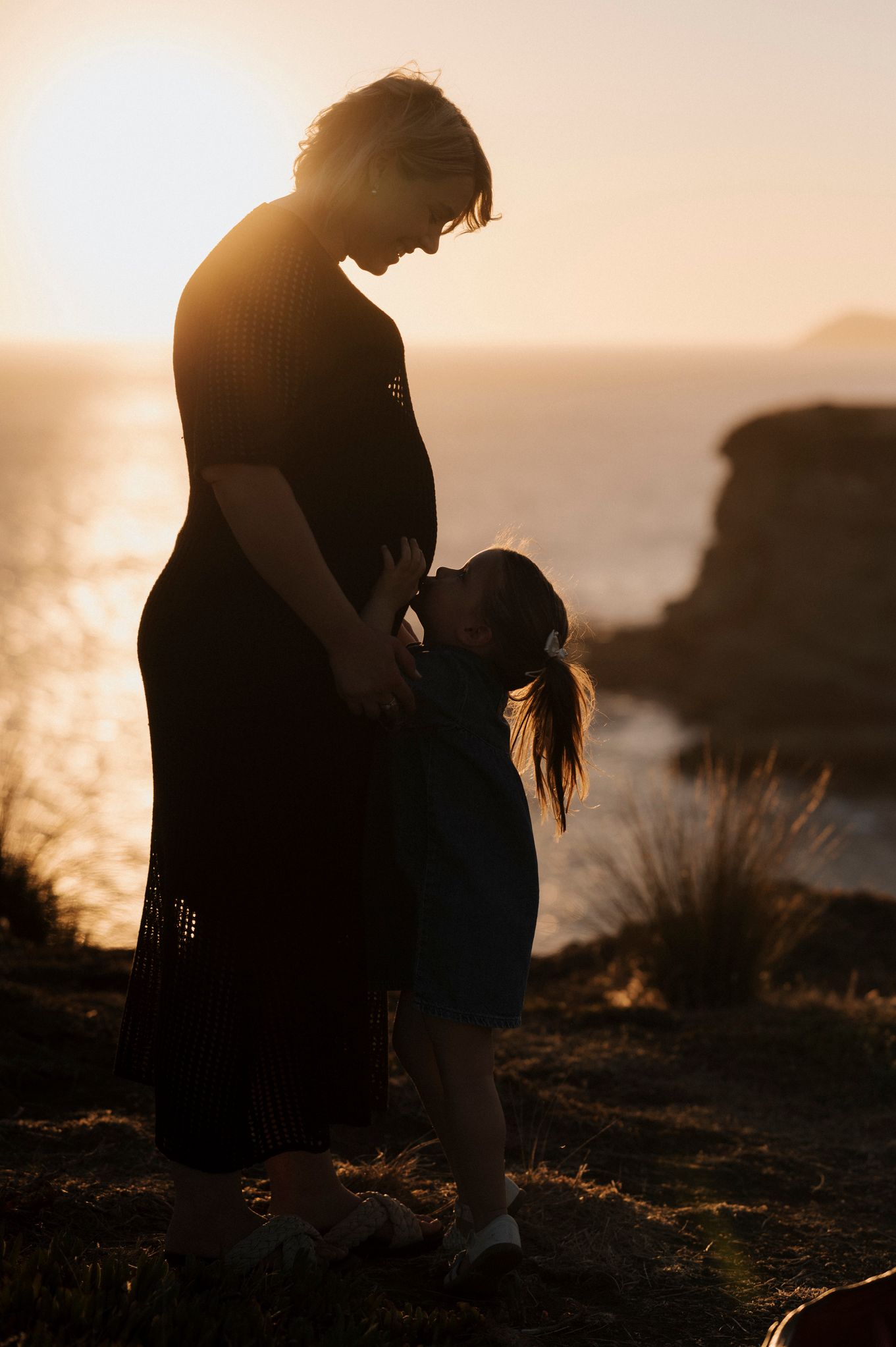 Silhouetted figures share a tender moment at sunset on a coastal clifftop with golden sunlight streaming through.