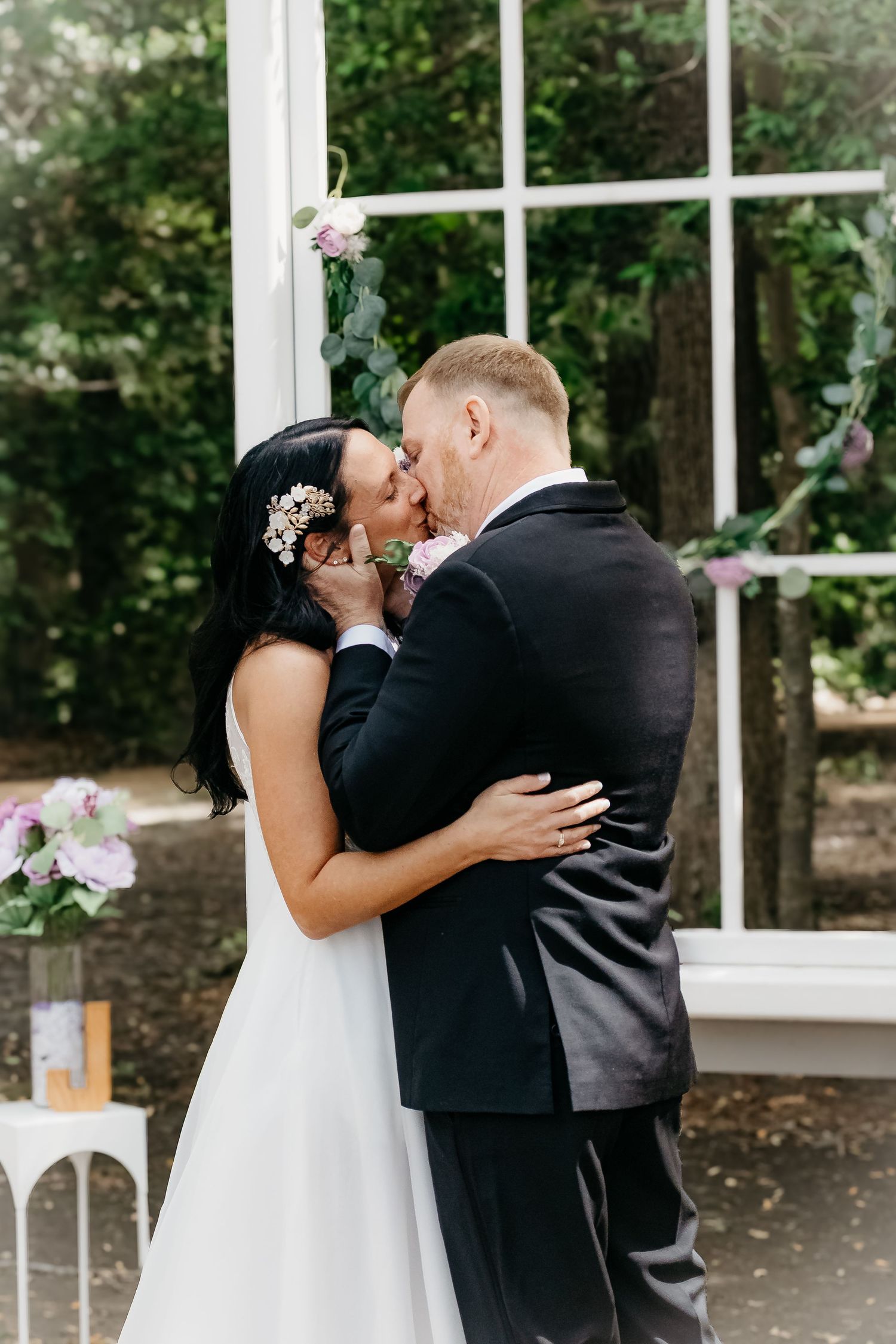 Couple shares a romantic kiss during their outdoor wedding ceremony near a white-framed window adorned with greenery.