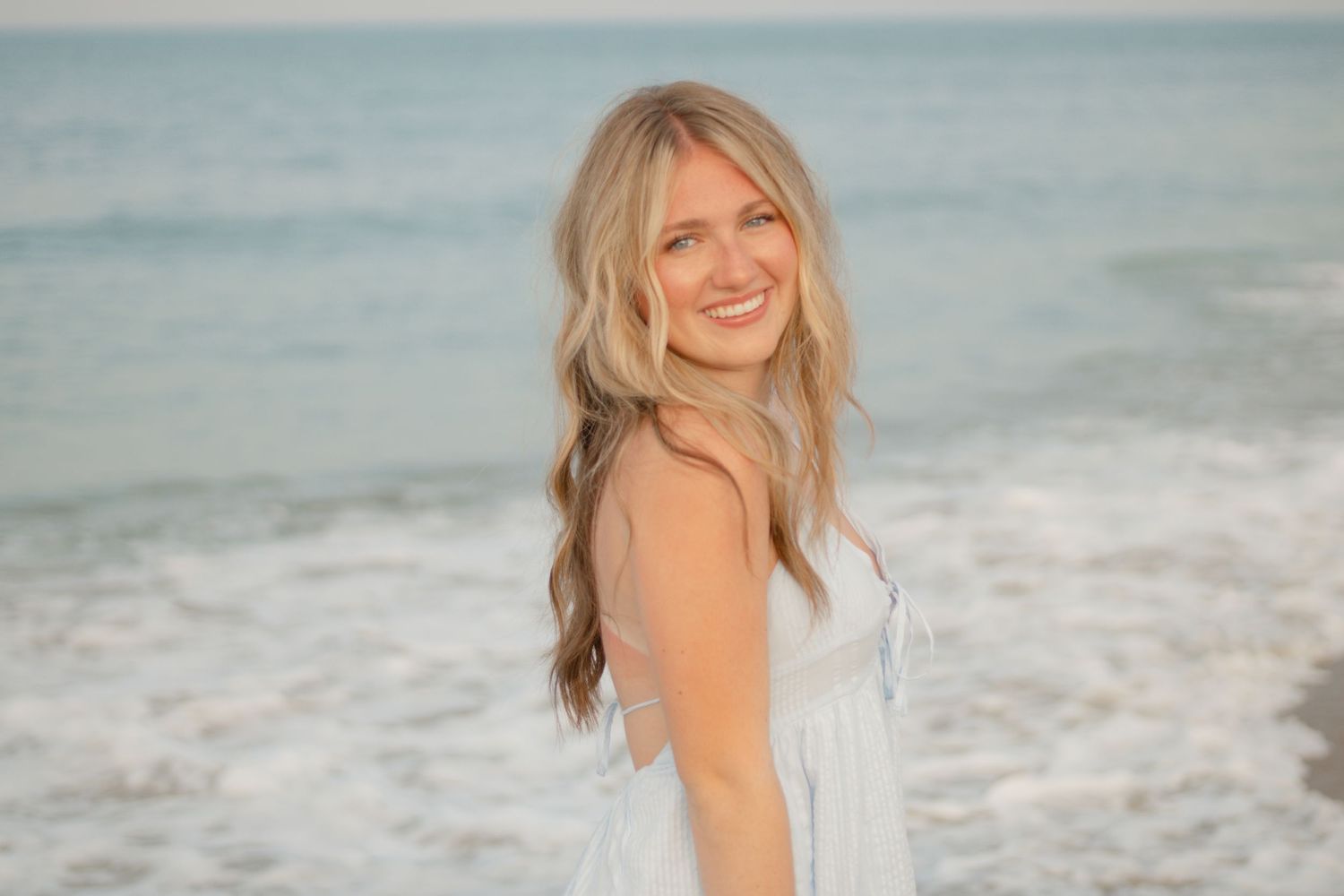 Woman in white dress posing on beach at sunset with ocean waves in background.
