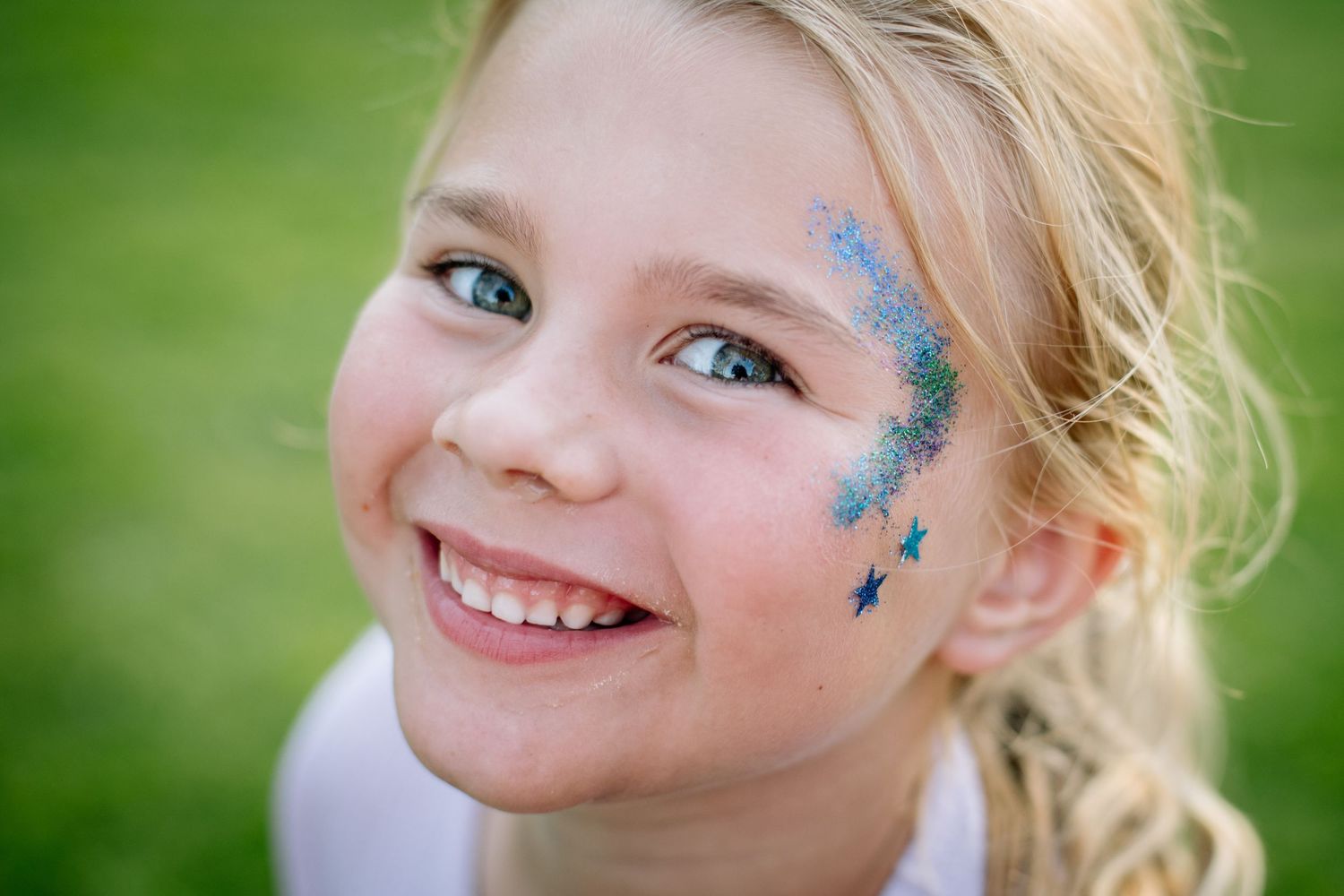 Young child with sparkly blue glitter face paint and a bright smile against a green outdoor background.