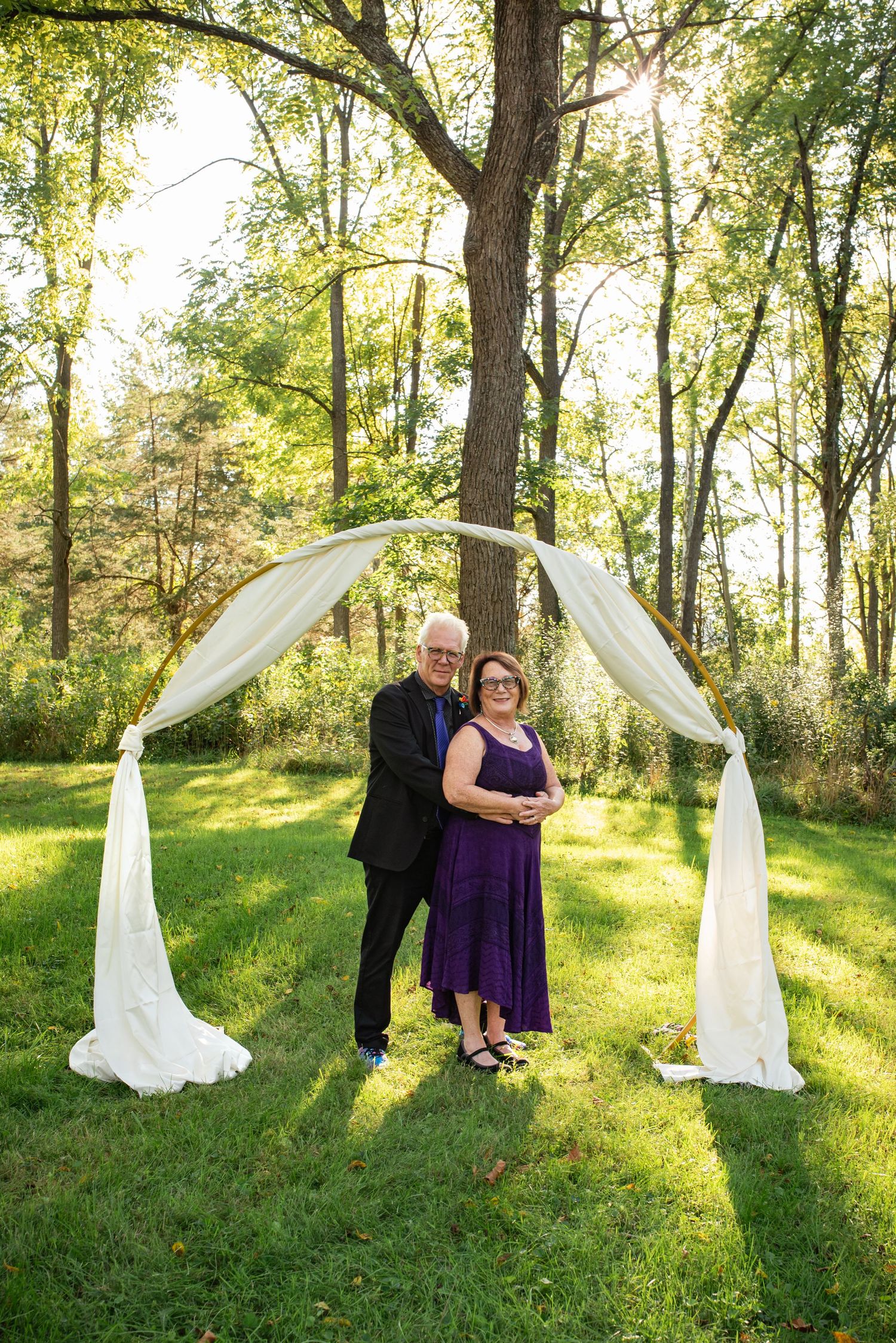Couple poses beneath flowing white fabric arch in sunlit woodland setting.