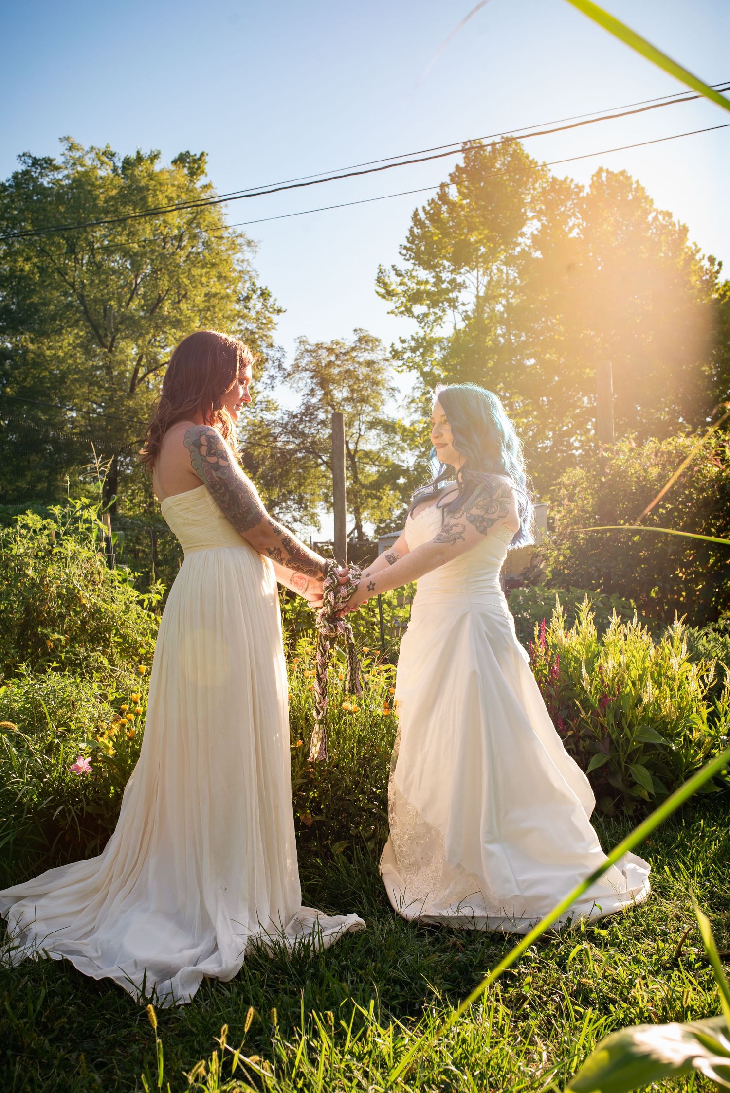 Two brides in white gowns hold hands in sunlit garden ceremony.