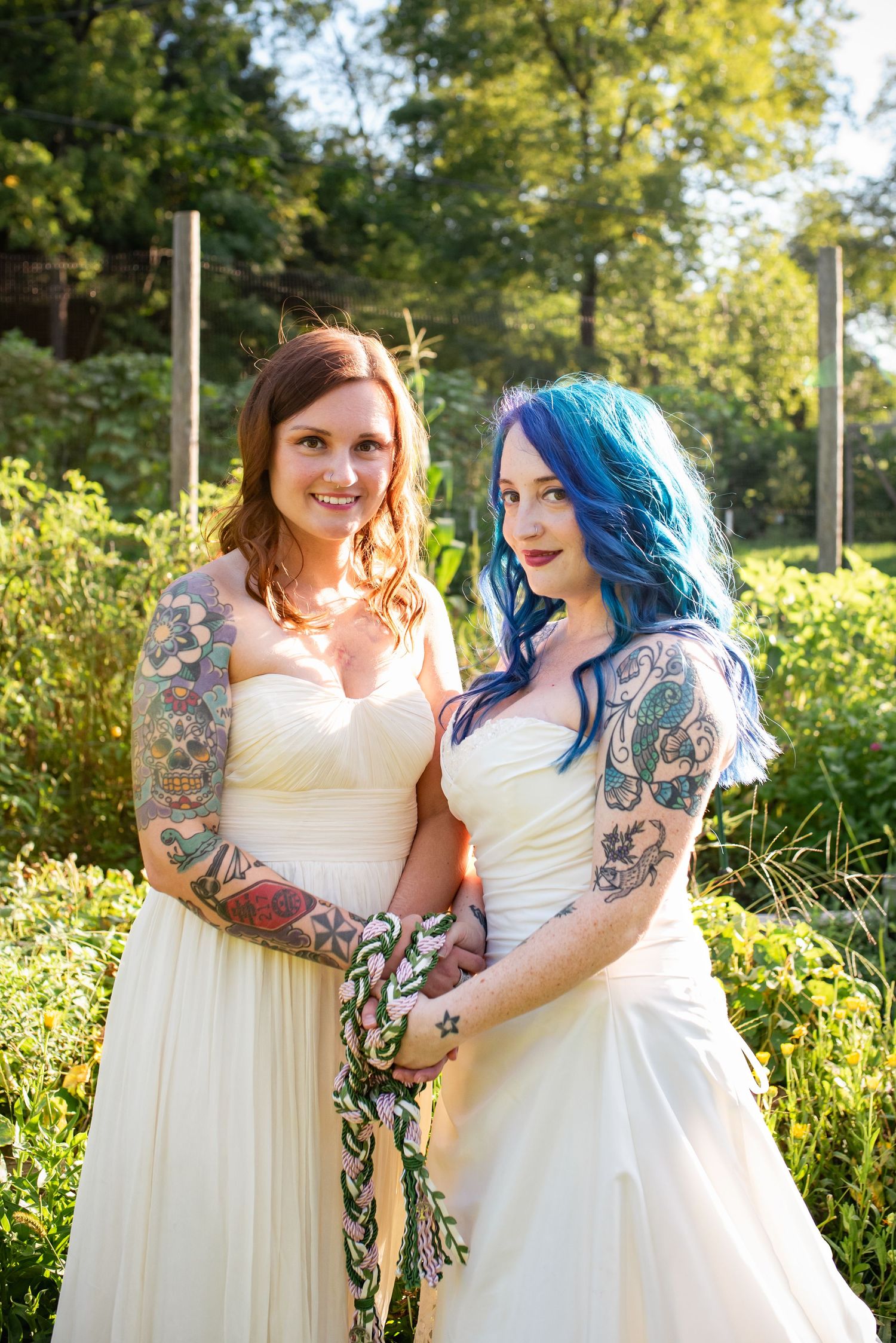 Two brides in white wedding dresses show off colorful tattoo sleeves in garden setting.