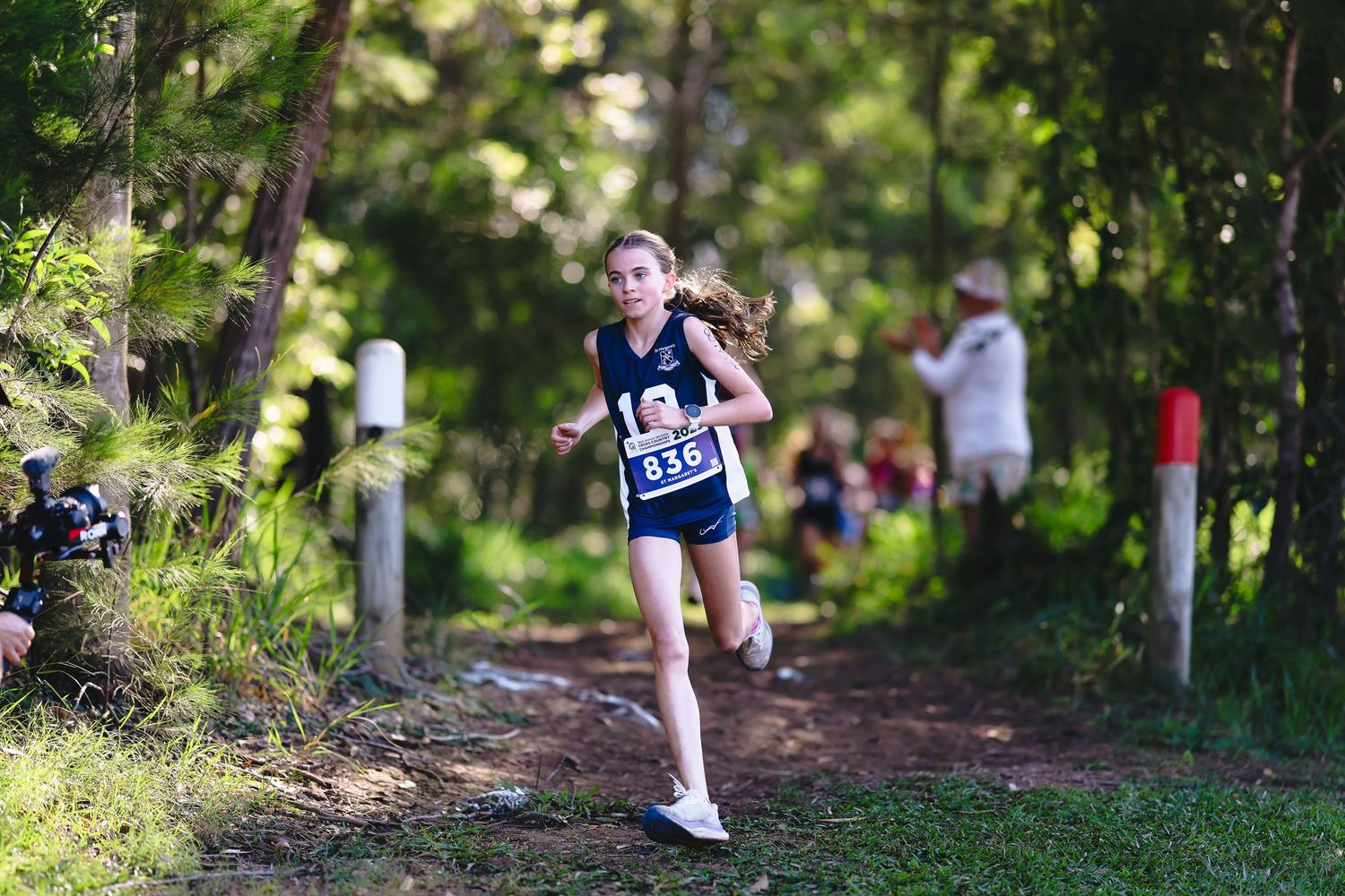 Runner in blue jersey competing at outdoor track meet with spectators and tents in background on sunny day.