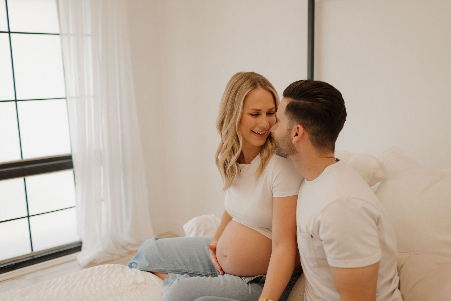 Intimate maternity photoshoot in bright minimalist room with white curtains.