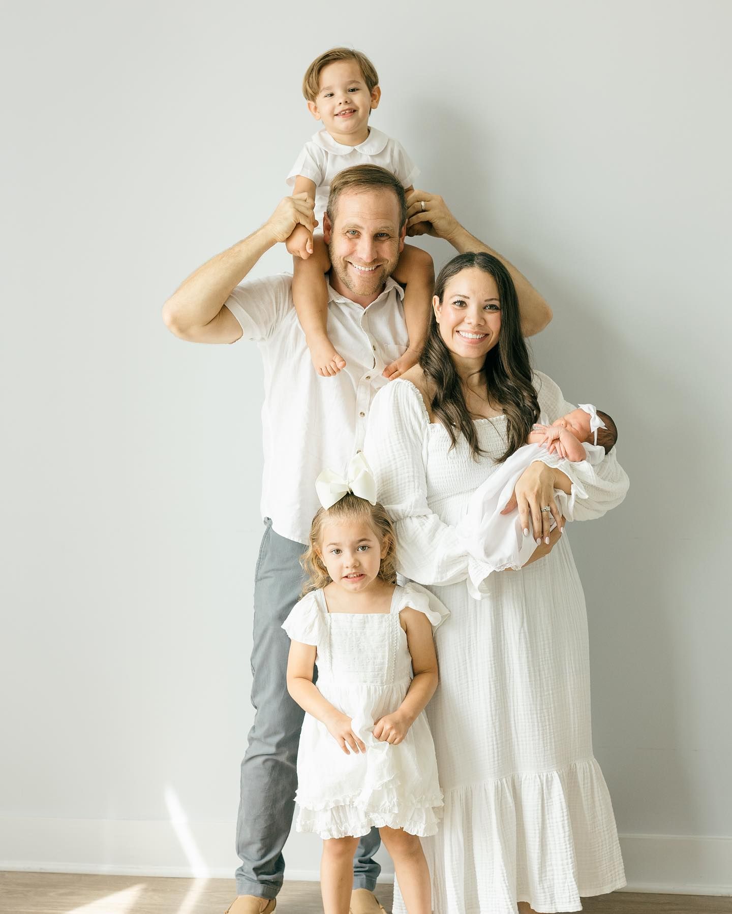 Contemporary family portrait with parents and children in coordinated white outfits against neutral background.