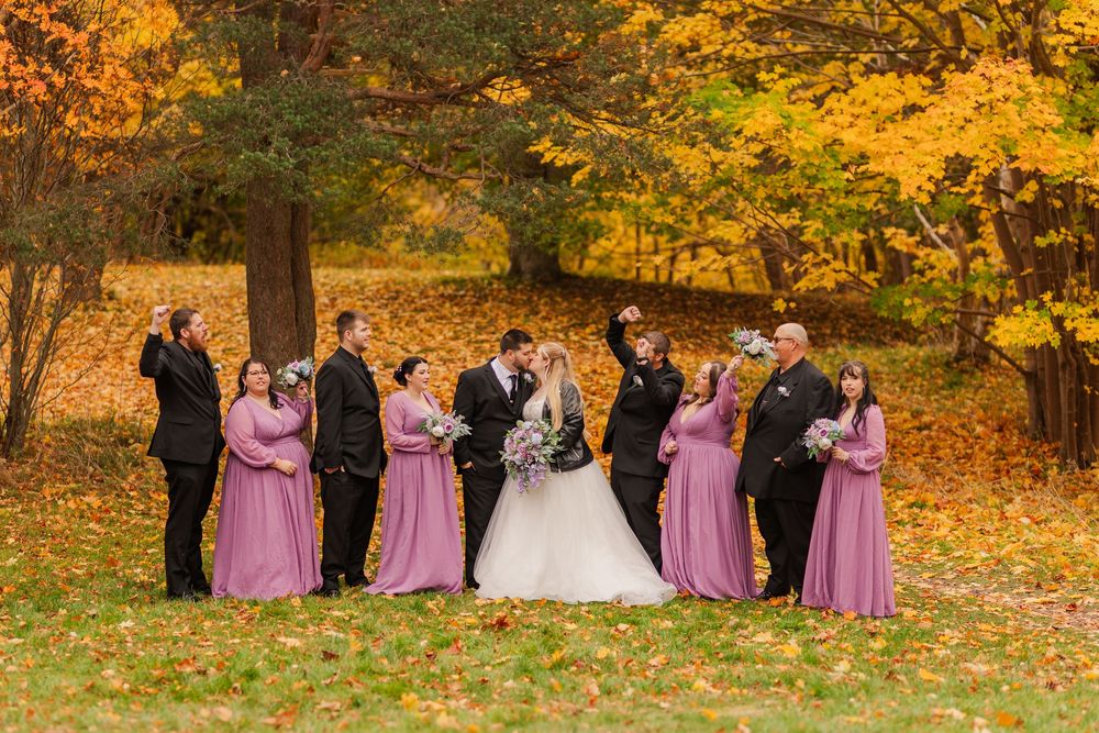 Bridal party in pink dresses stands together against a backdrop of golden autumn leaves in a park.