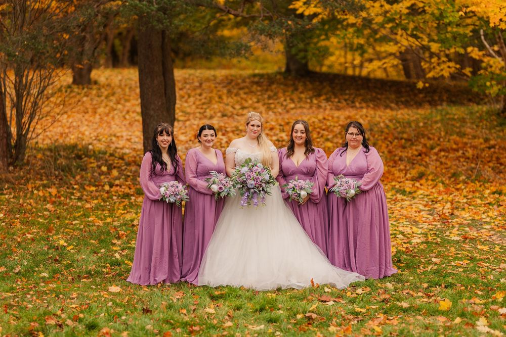 Bridesmaids in flowing mauve gowns hold bouquets against a backdrop of golden fall leaves.