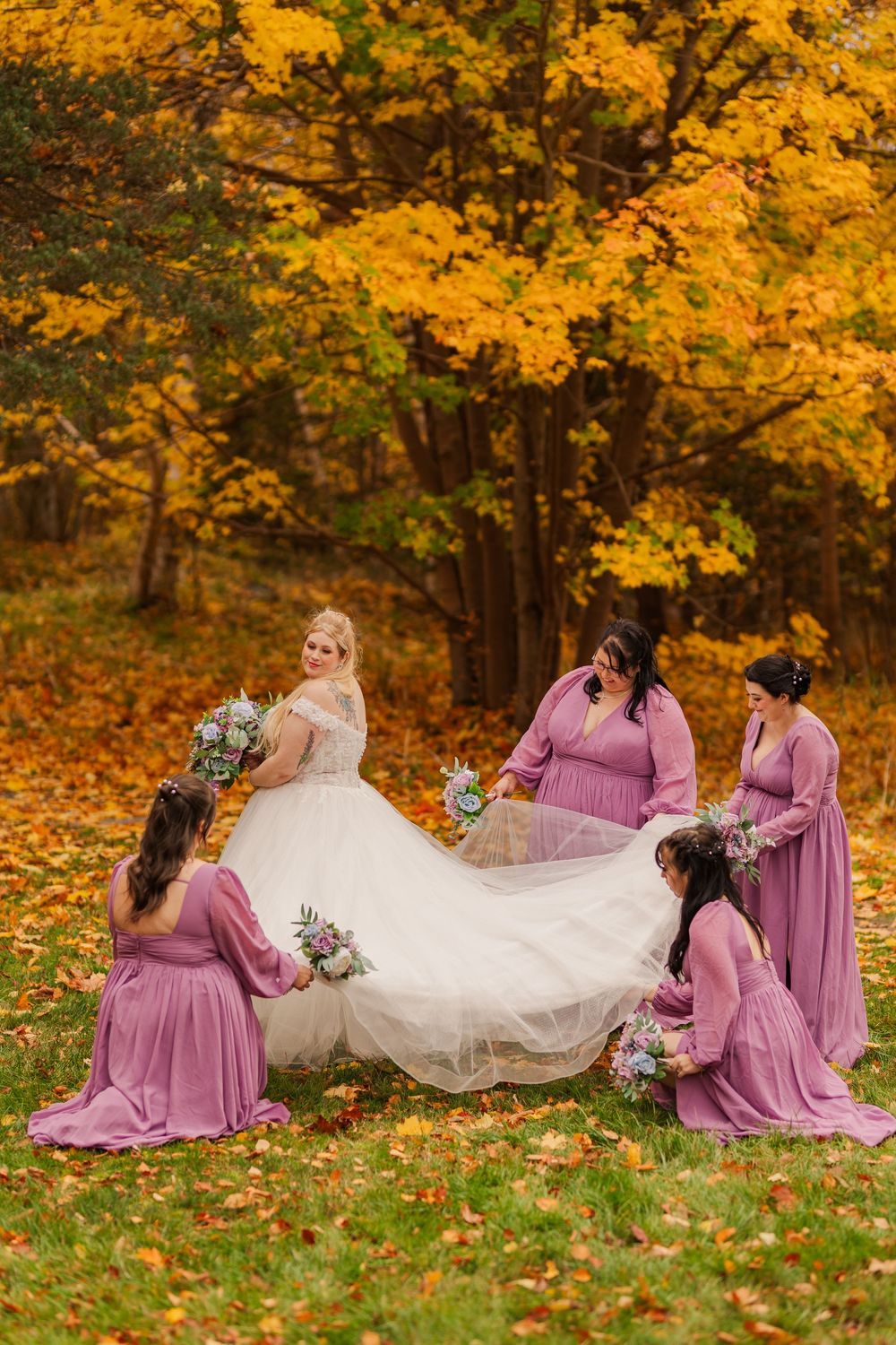 A wedding party in mauve bridesmaid dresses poses playfully on autumn leaves with flowing white bridal gown.