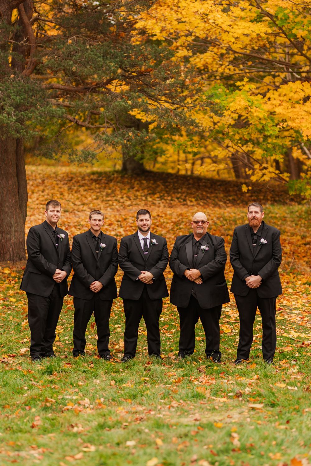 Groomsmen in black suits stand together on fallen autumn leaves under golden trees.