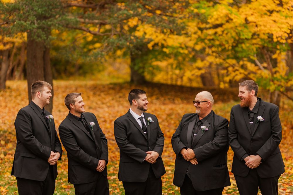 Wedding party members in dark suits share a candid moment against vibrant autumn foliage in the background.