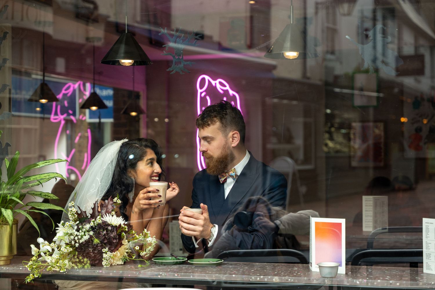 A couple shares an intimate moment in front of a restaurant window with neon lights and floral decorations.