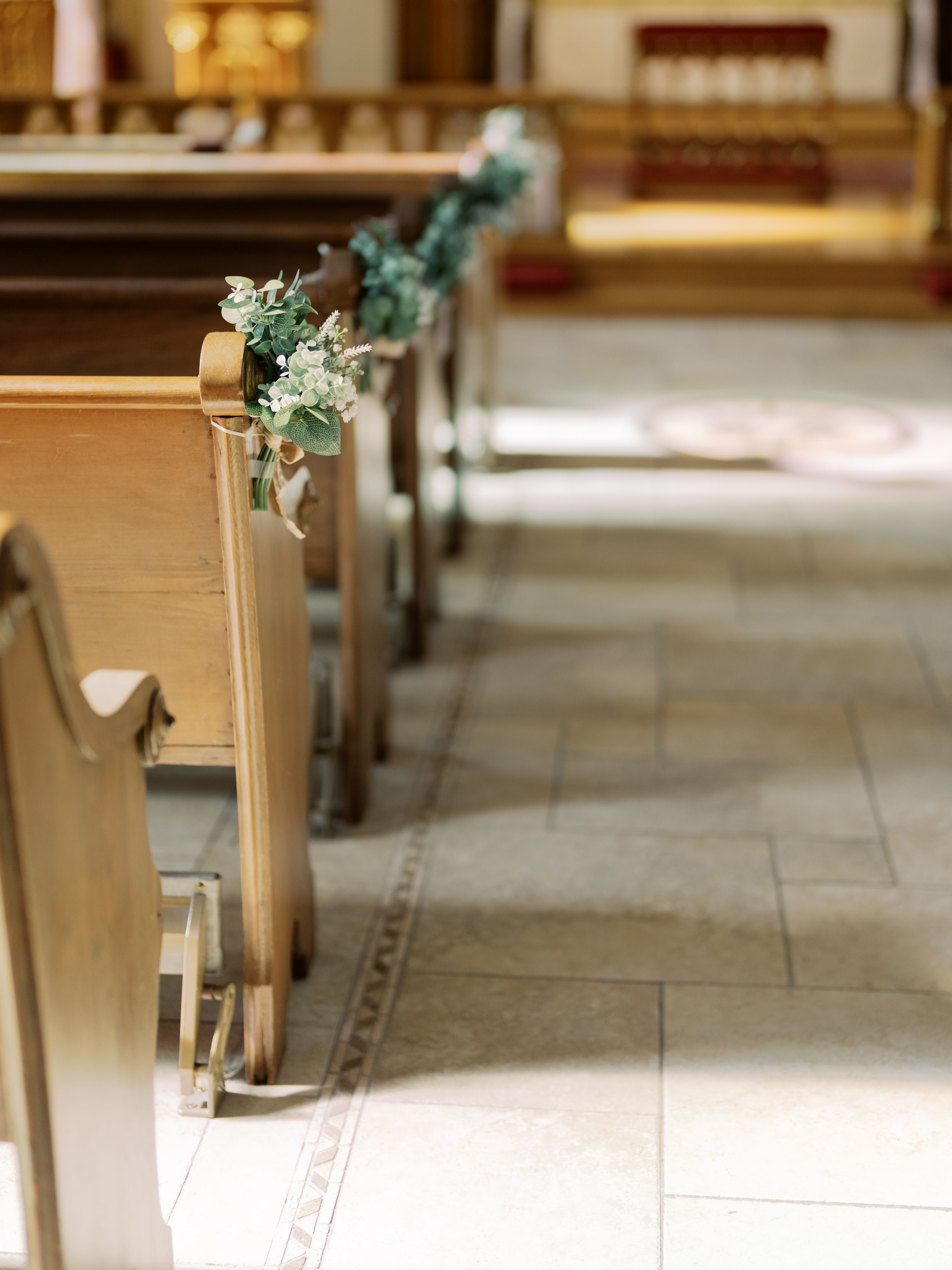 Wooden church pews decorated with delicate greenery and white floral arrangements line the aisle for a wedding ceremony.
