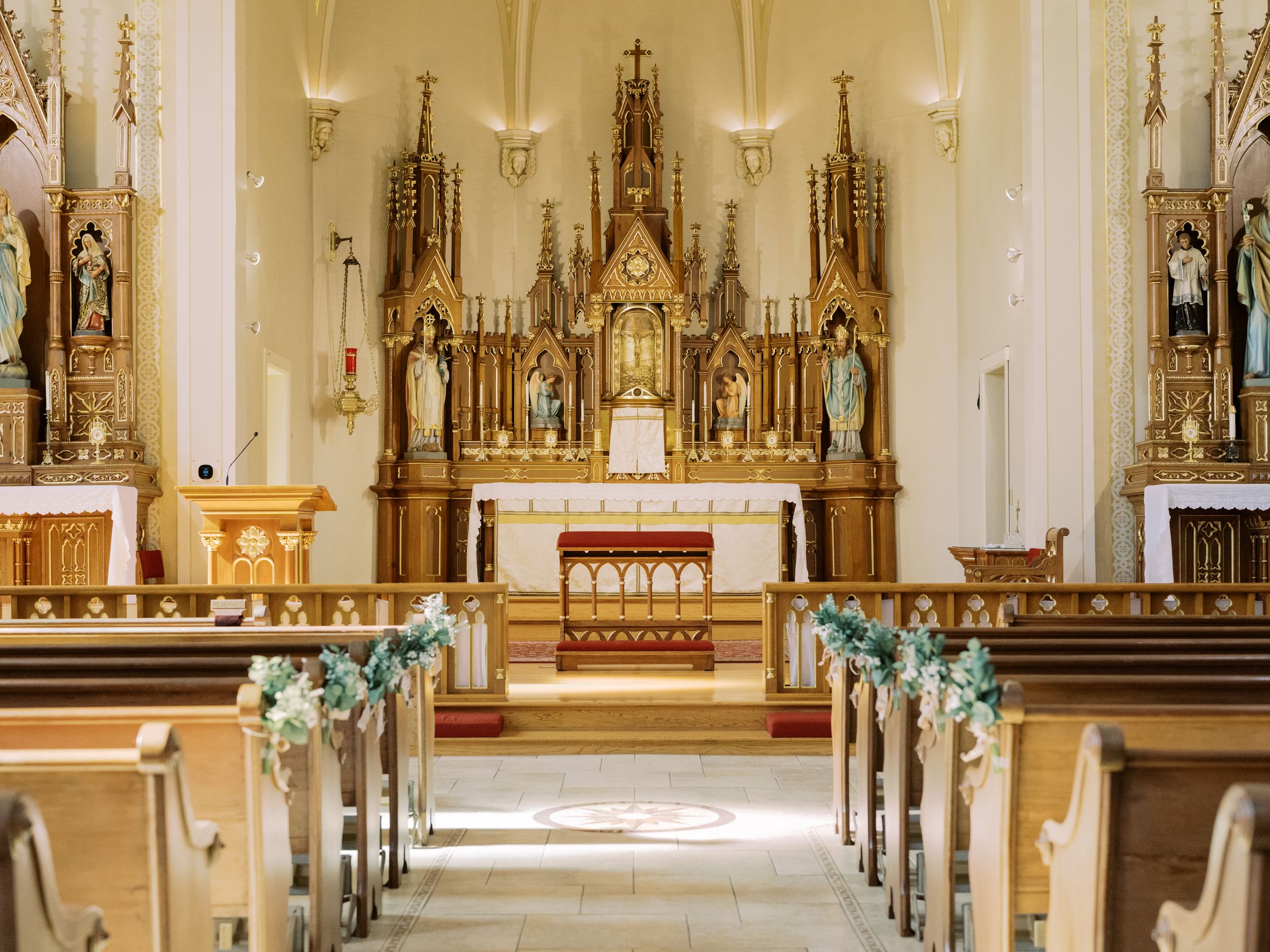 Interior view of a traditional Catholic church with ornate altar, wooden pews, and elegant chandeliers lining the aisle.