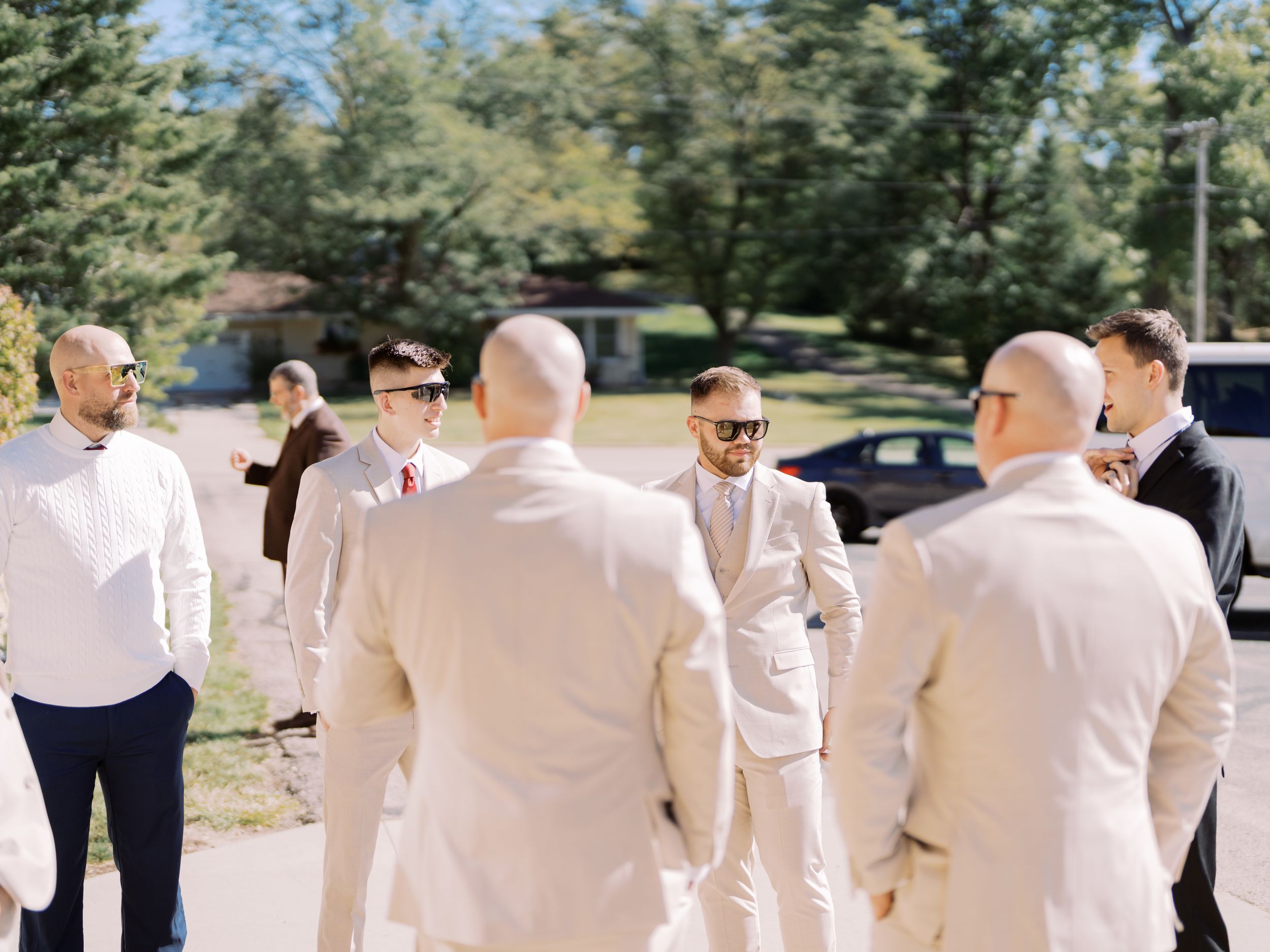 Wedding party members in light colored suits stand together outdoors on a sunny day in a garden setting.