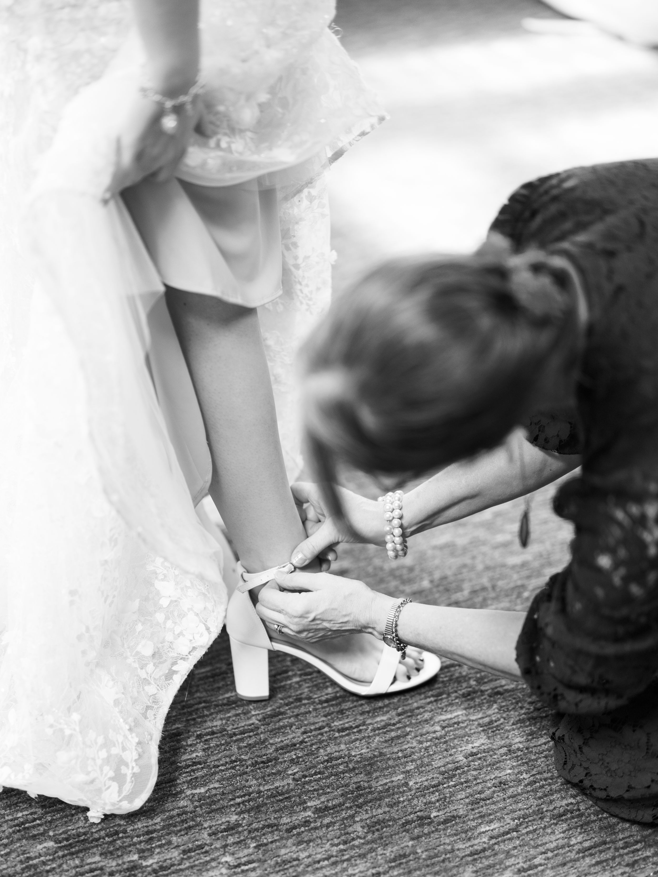 Black and white photo showing someone adjusting a bride's delicate white sandal during wedding preparations.