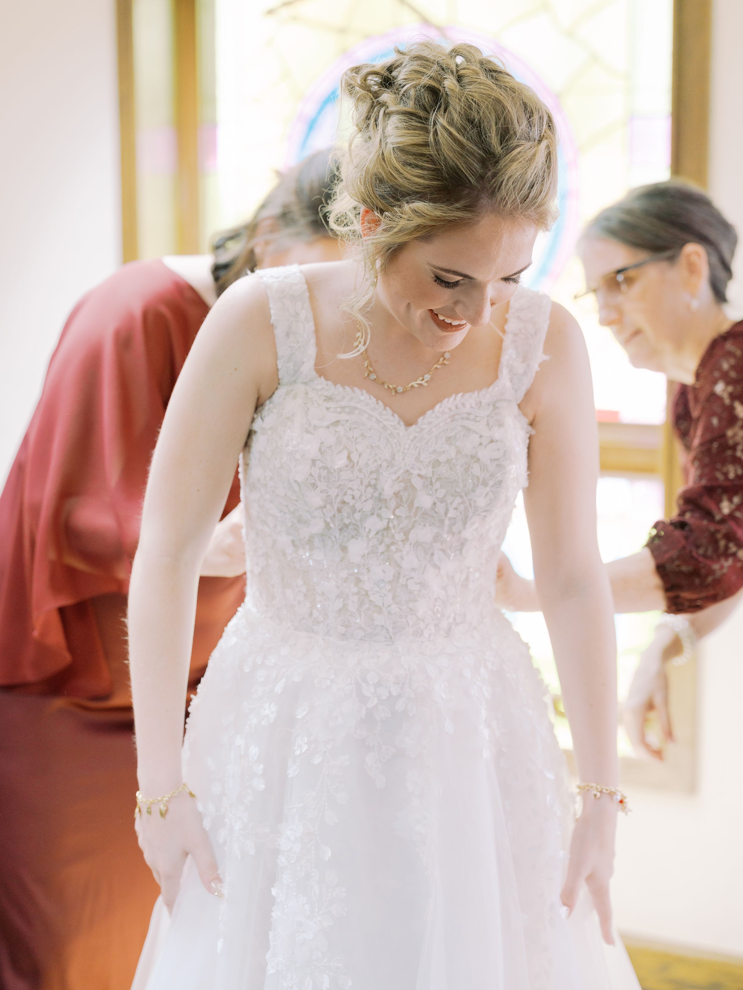 A bride in a white lace wedding dress gets help with final adjustments during wedding preparations.