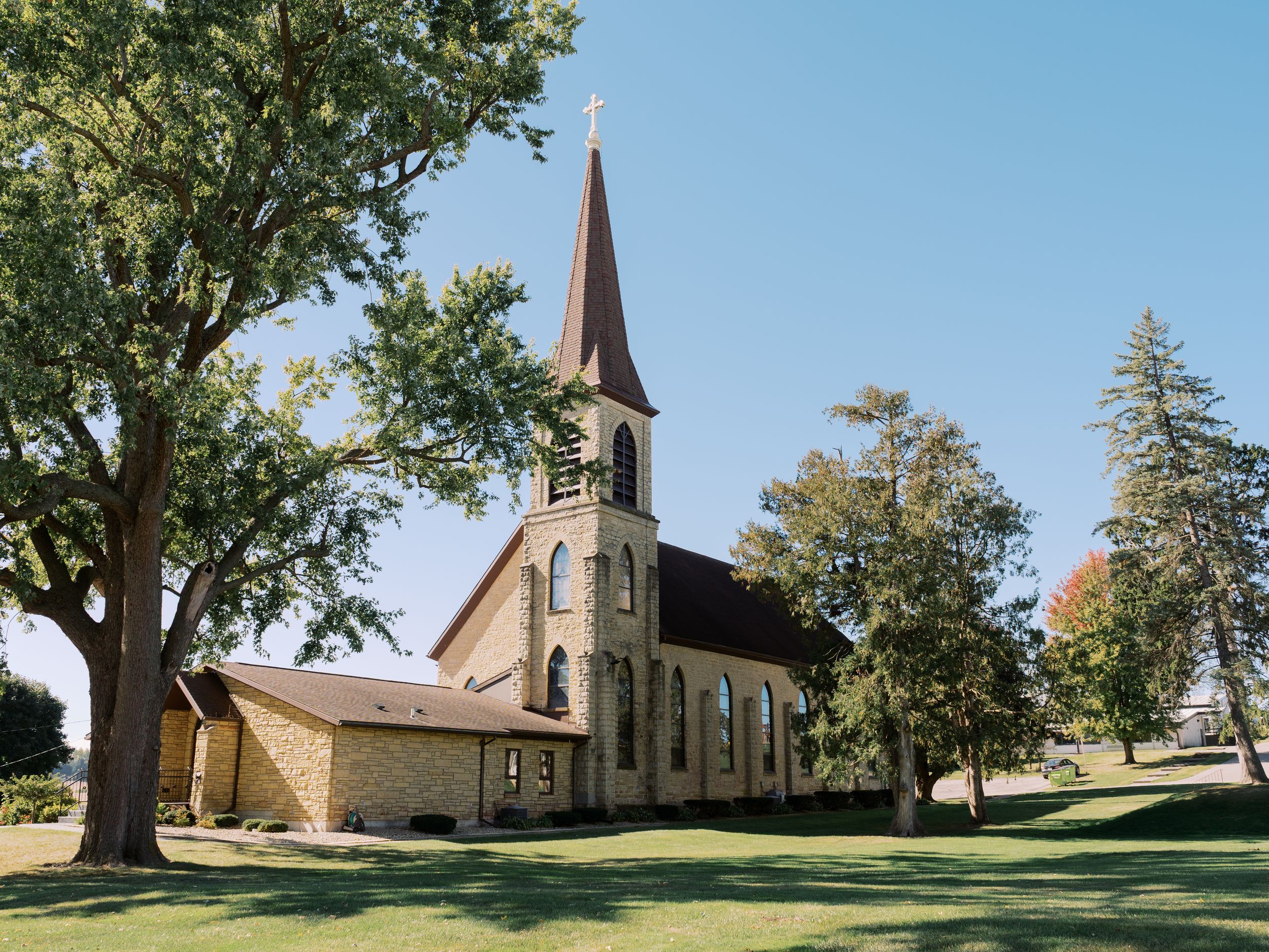Historic church with tall spire stands on a grassy hill surrounded by trees in a rural countryside setting.