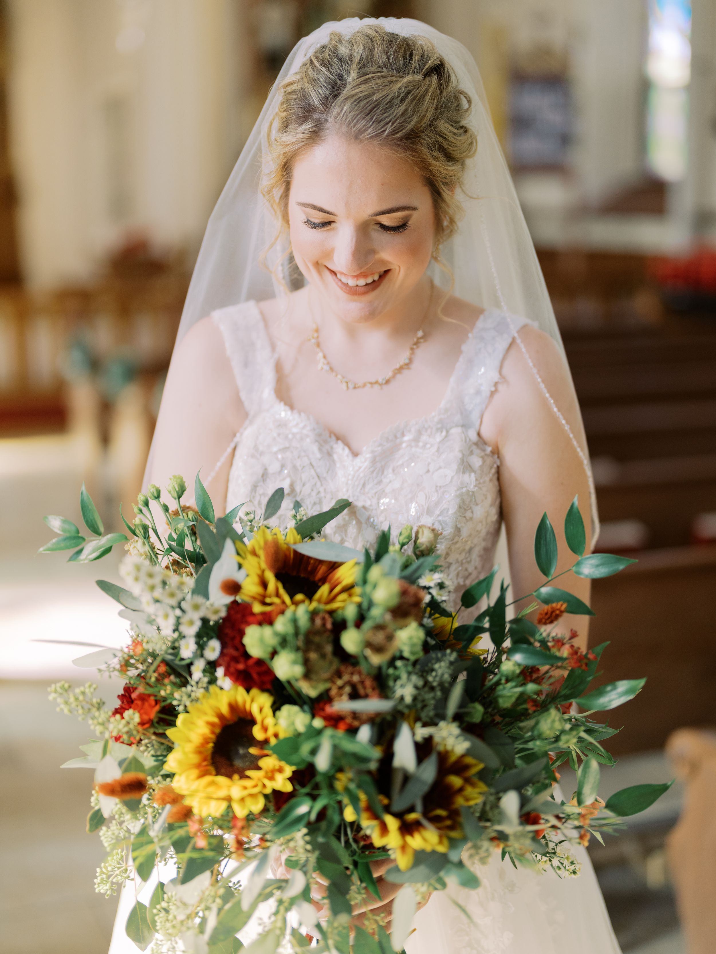 A bride in a white lace dress holds a vibrant bouquet featuring sunflowers, greenery and fall-colored blooms.