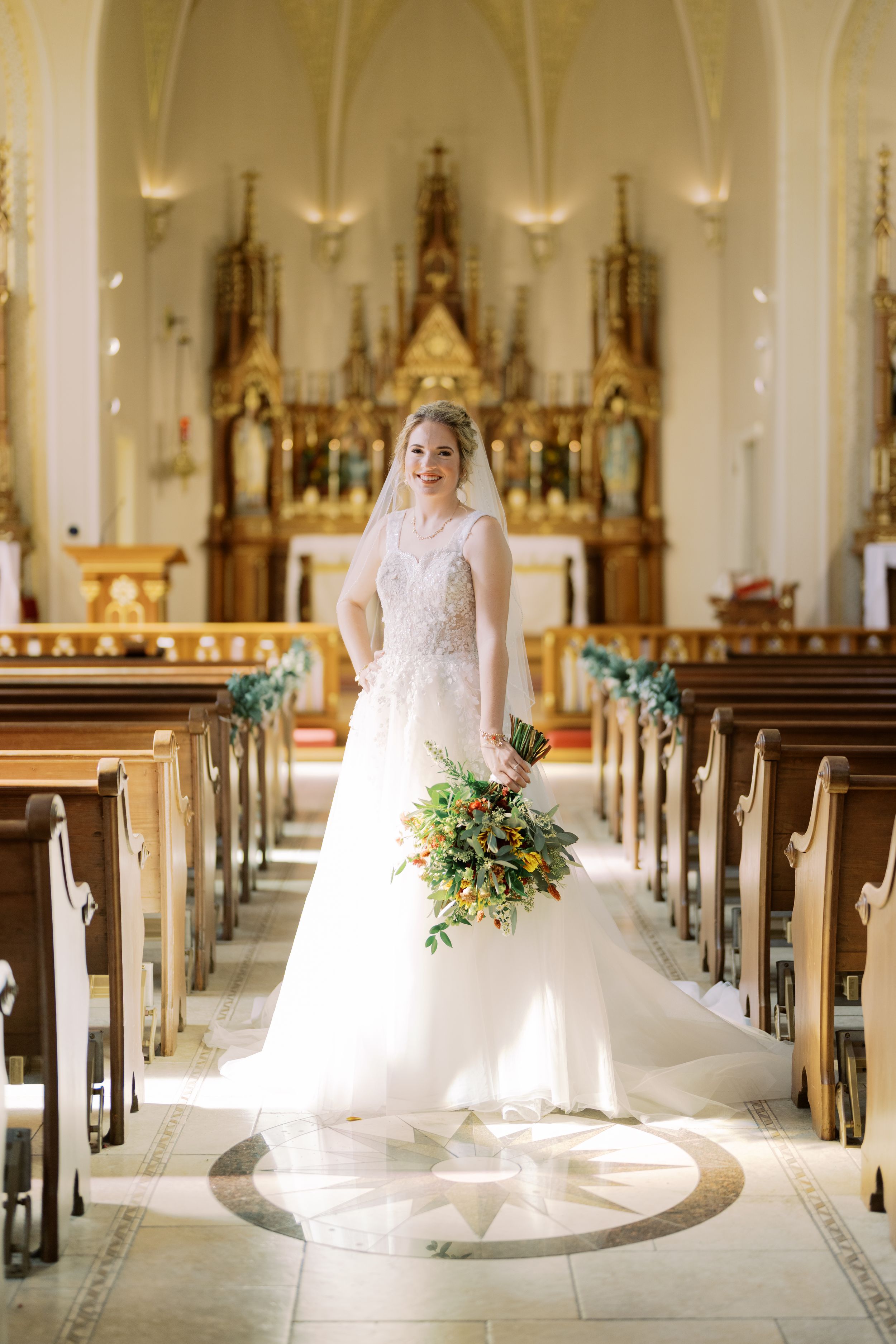 Sunlit cathedral aisle with white pews and ornate altar, featuring a bride in a flowing lace gown holding yellow bouquet.