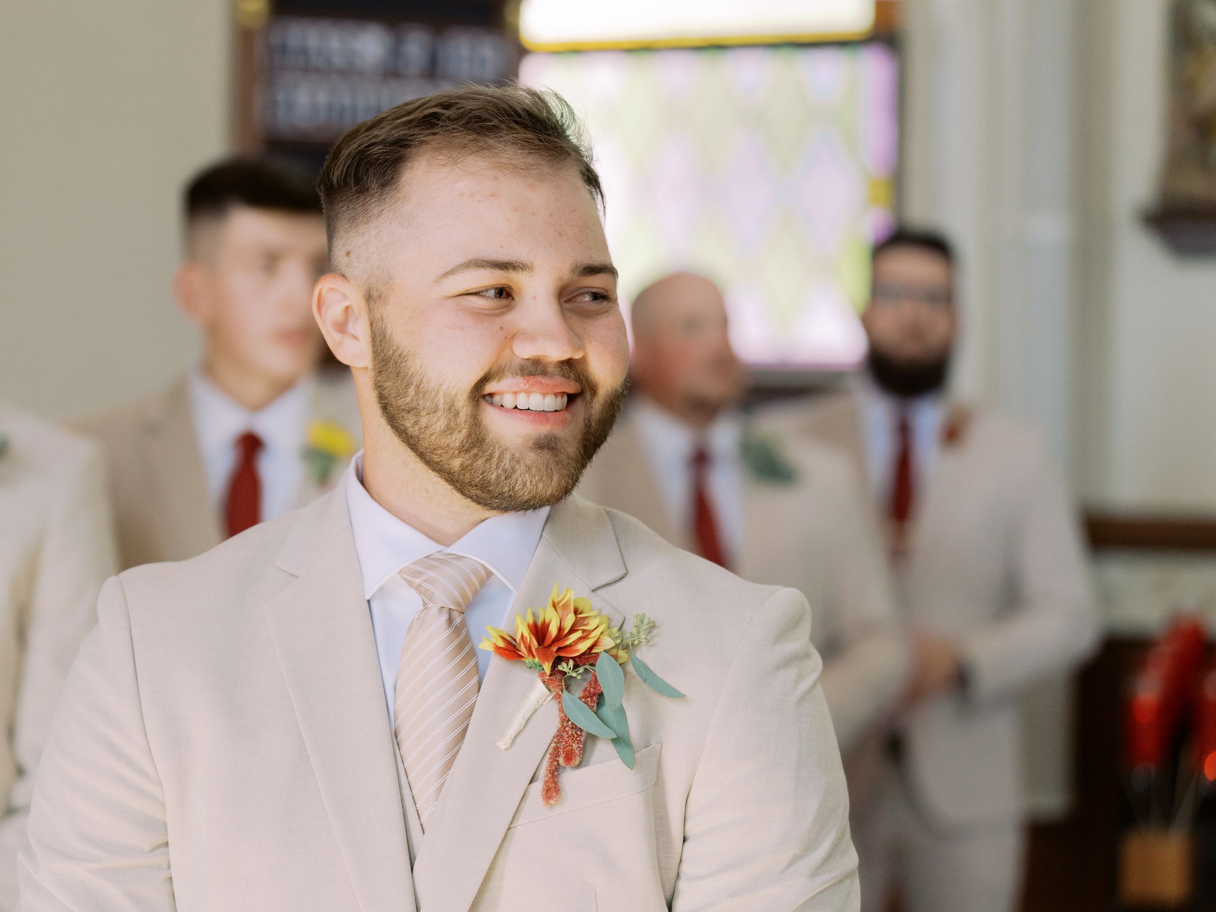 Groomsmen in light beige suits with colorful boutonnieres smile during a wedding celebration.