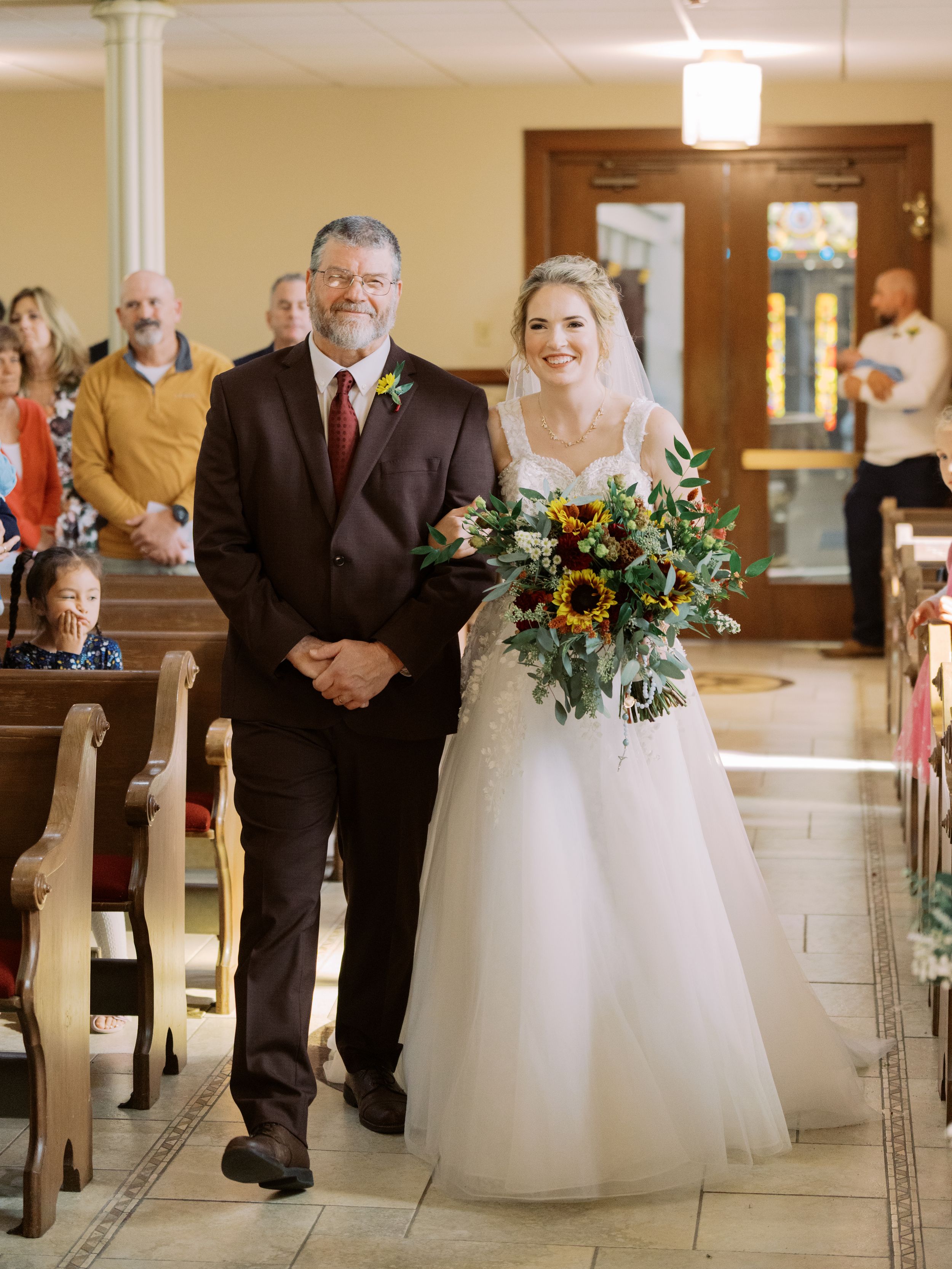 A wedding ceremony in progress inside a church with floral arrangements on white pedestals along the aisle.