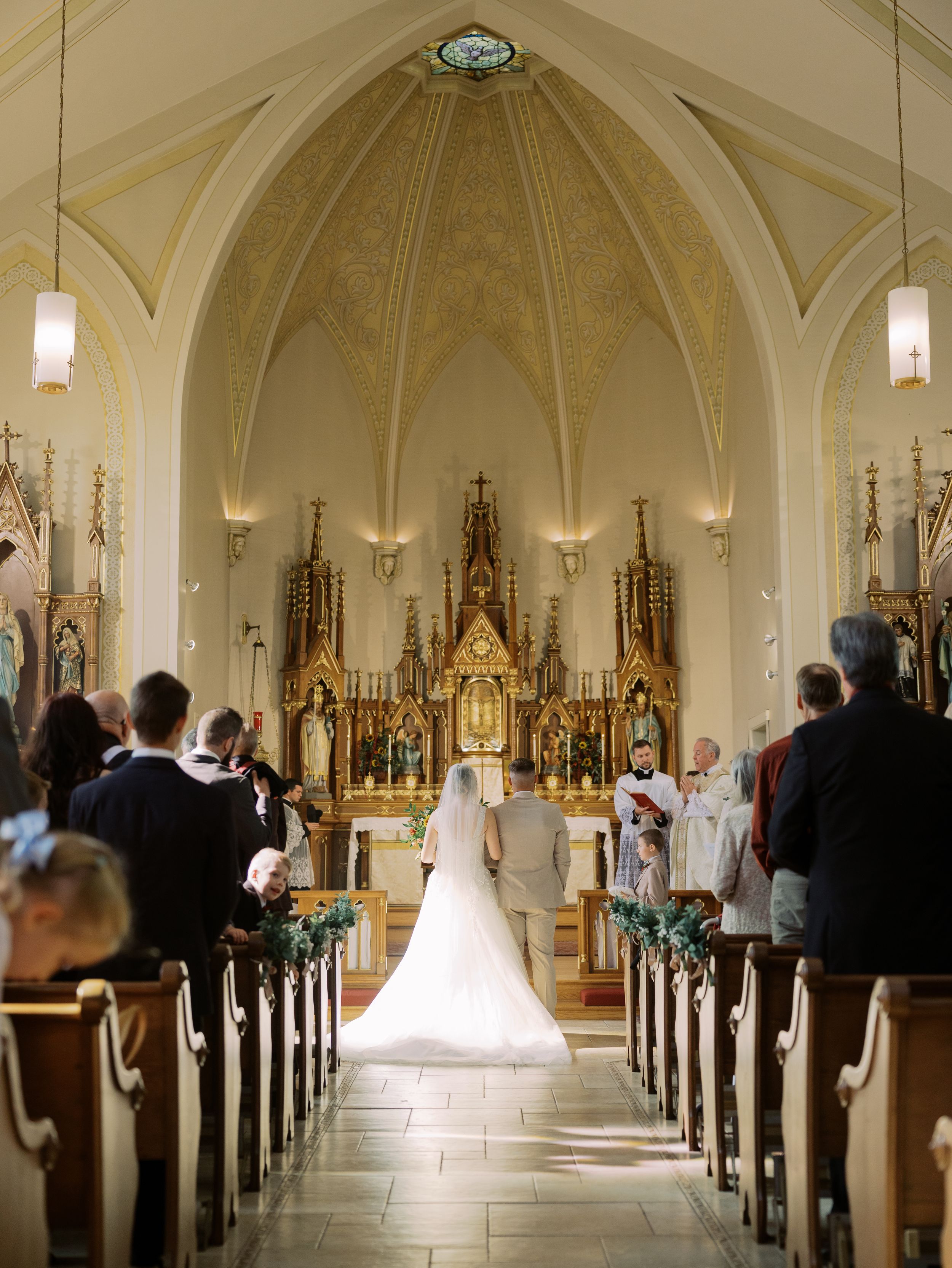 A panoramic view of a traditional Catholic church interior during a wedding ceremony with white decor and ornate altars.