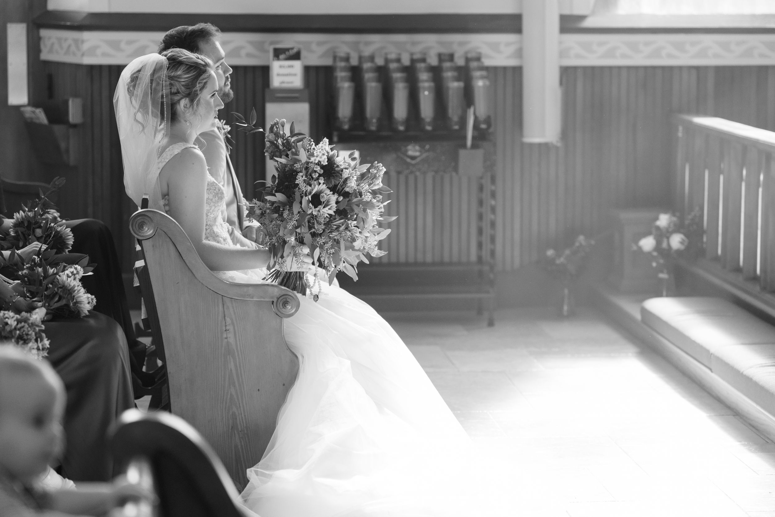 A bride in a flowing white gown holds a bouquet while standing in soft window light at a church venue.