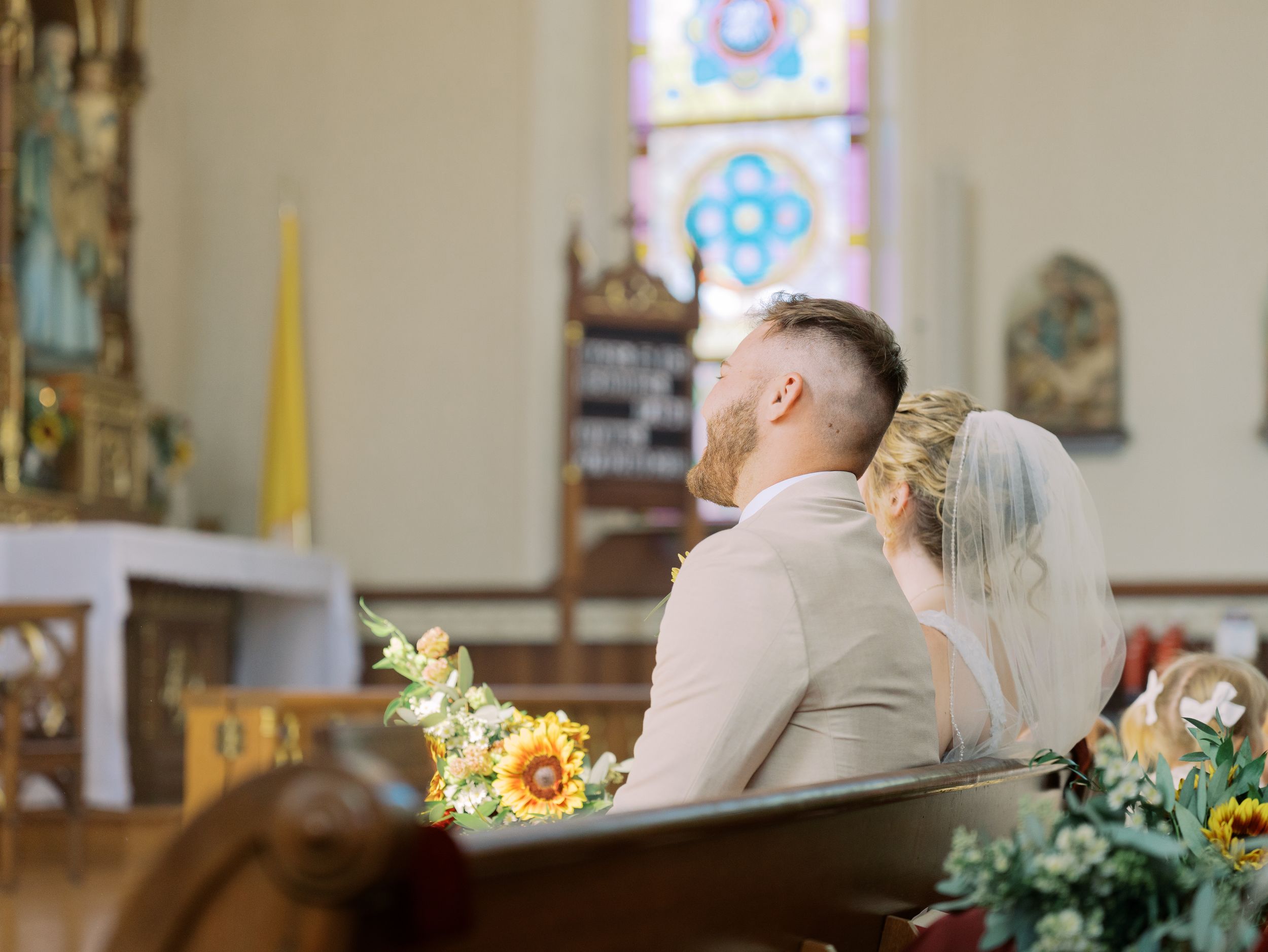 A couple sits in a church pew decorated with yellow sunflowers during a wedding ceremony.