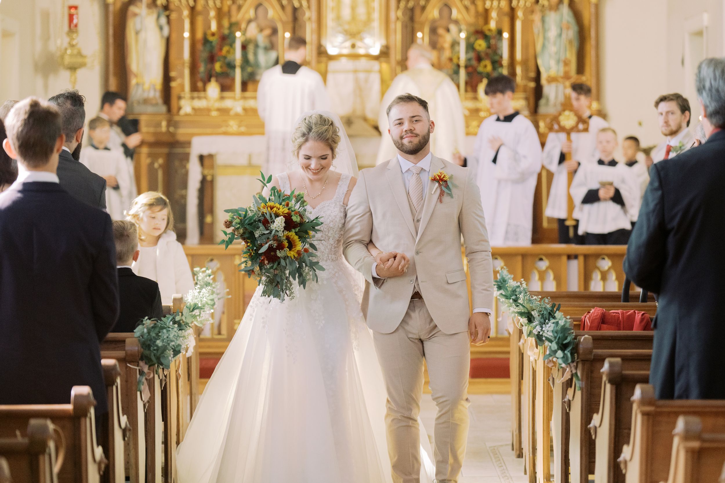 A wedding ceremony takes place in an ornate church with gold details and decorative altar as guests observe from wooden pews.