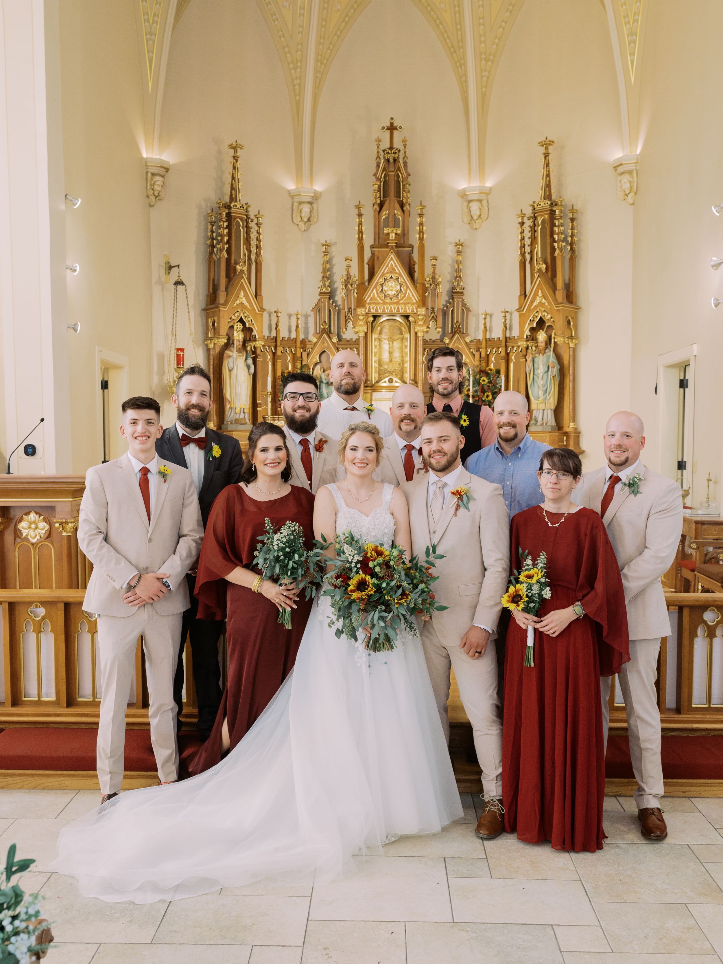 Wedding party poses in church sanctuary with burgundy bridesmaids dresses and tan groomsmen suits beside altar.