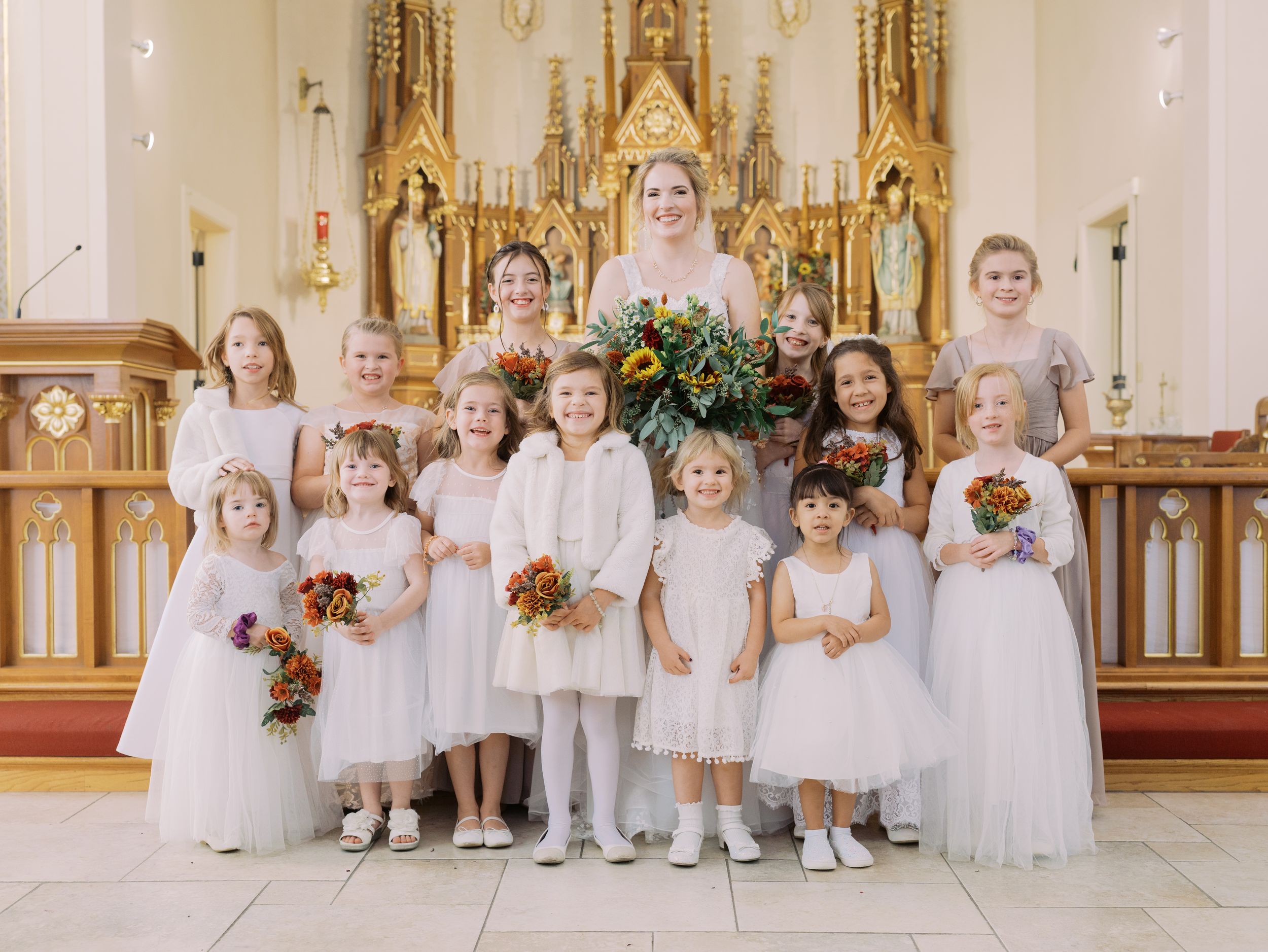 A row of flower girls in white dresses sit on church steps holding floral bouquets during a wedding ceremony.