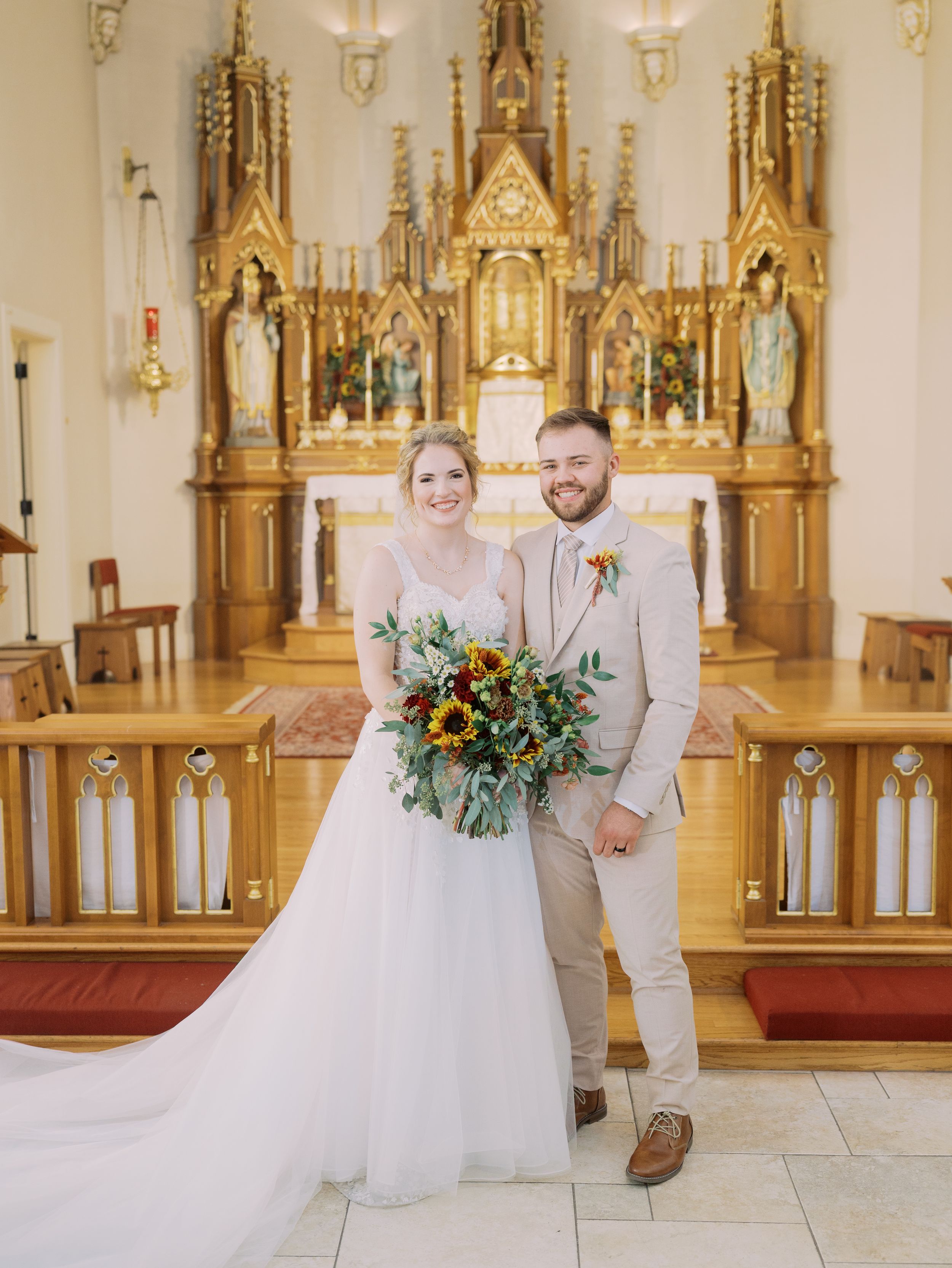 A couple in wedding attire stands together in an ornate church with golden gothic altars and white marble floors.