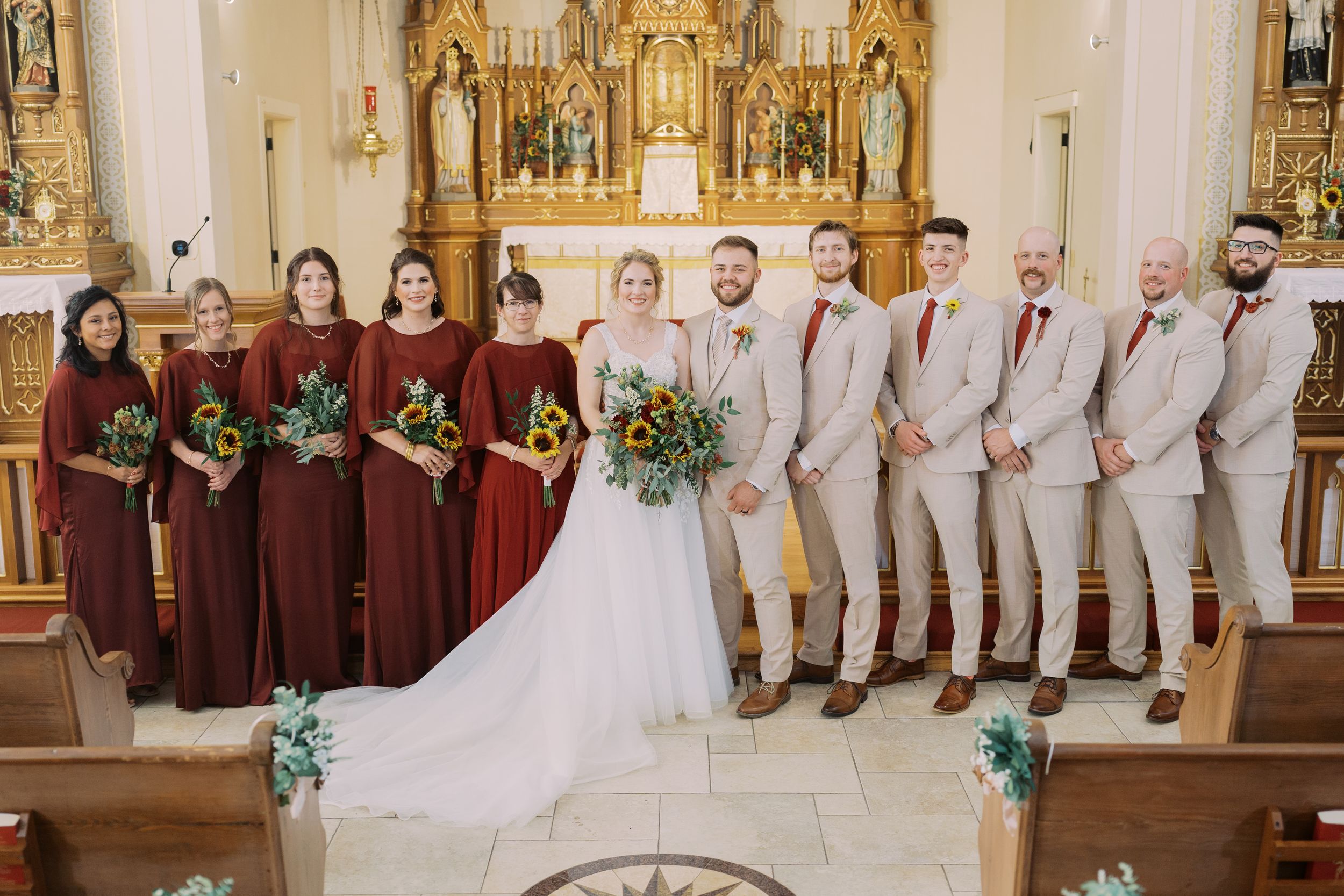 Wedding party members in burgundy dresses and gray suits pose together in a traditional church setting.
